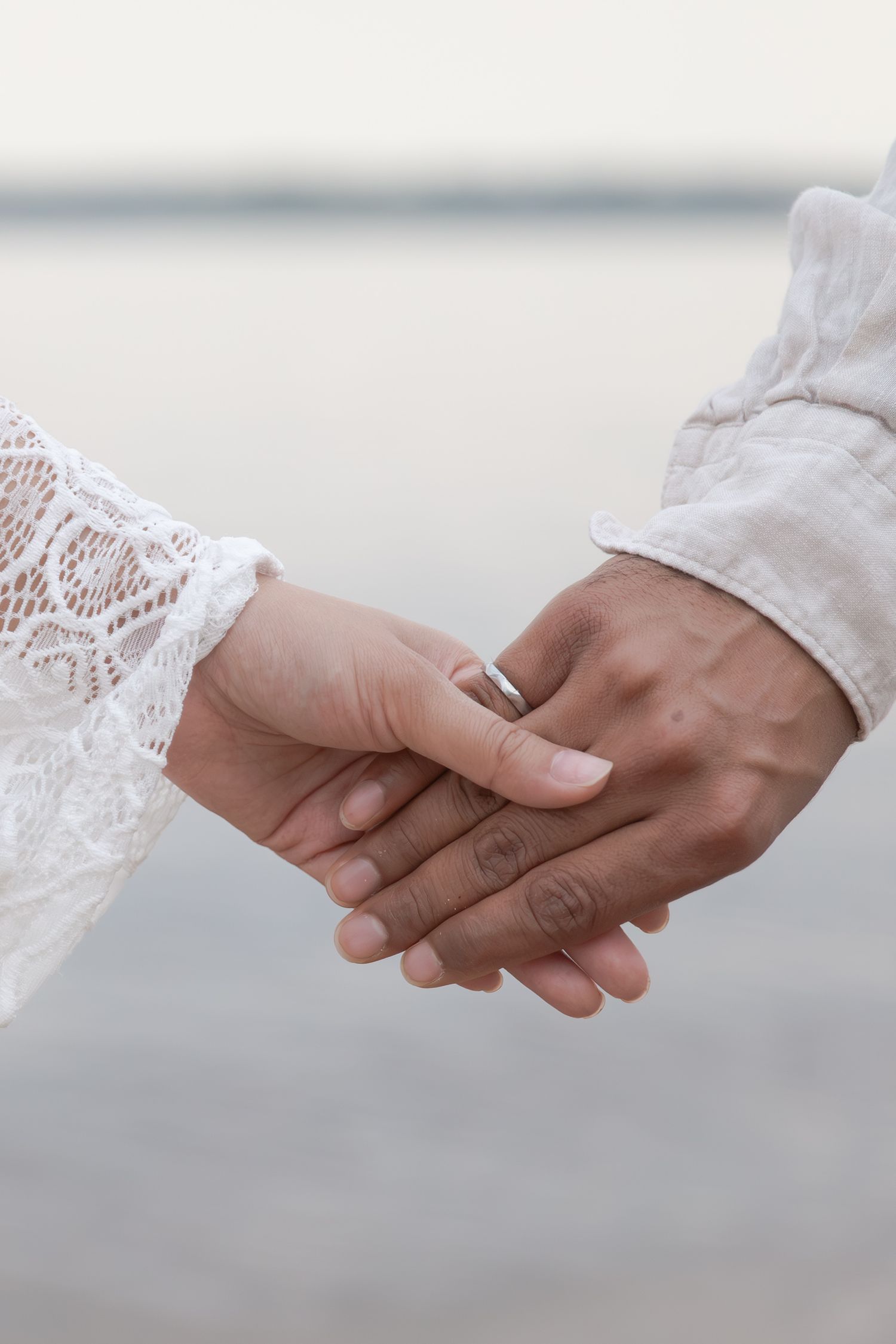 Two hands tenderly clasped together, one wearing delicate white lace and a wedding ring, against a soft blurred background.