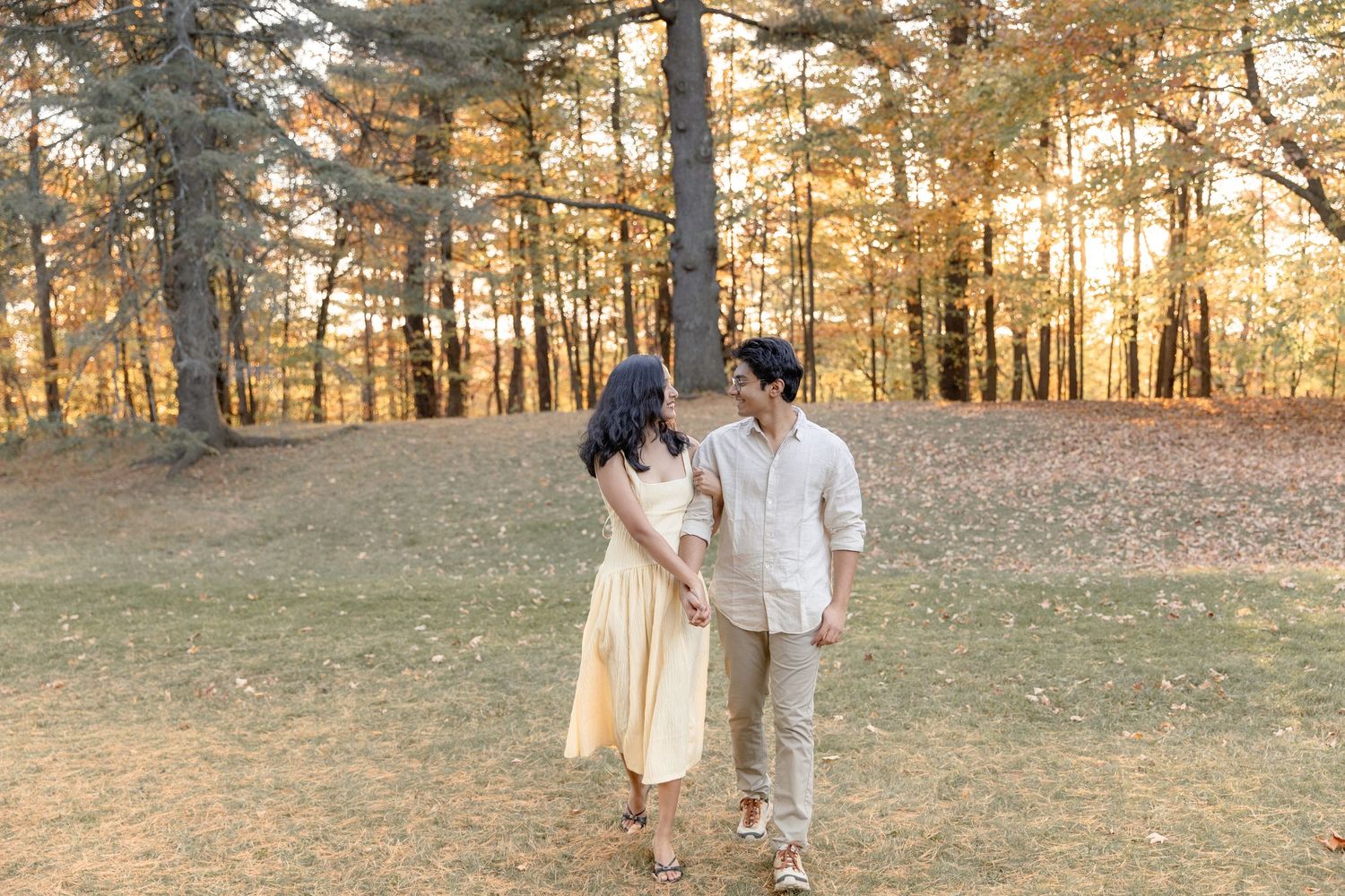 Couple walking hand in hand through a sunlit forest during golden hour as warm light filters through pine trees.