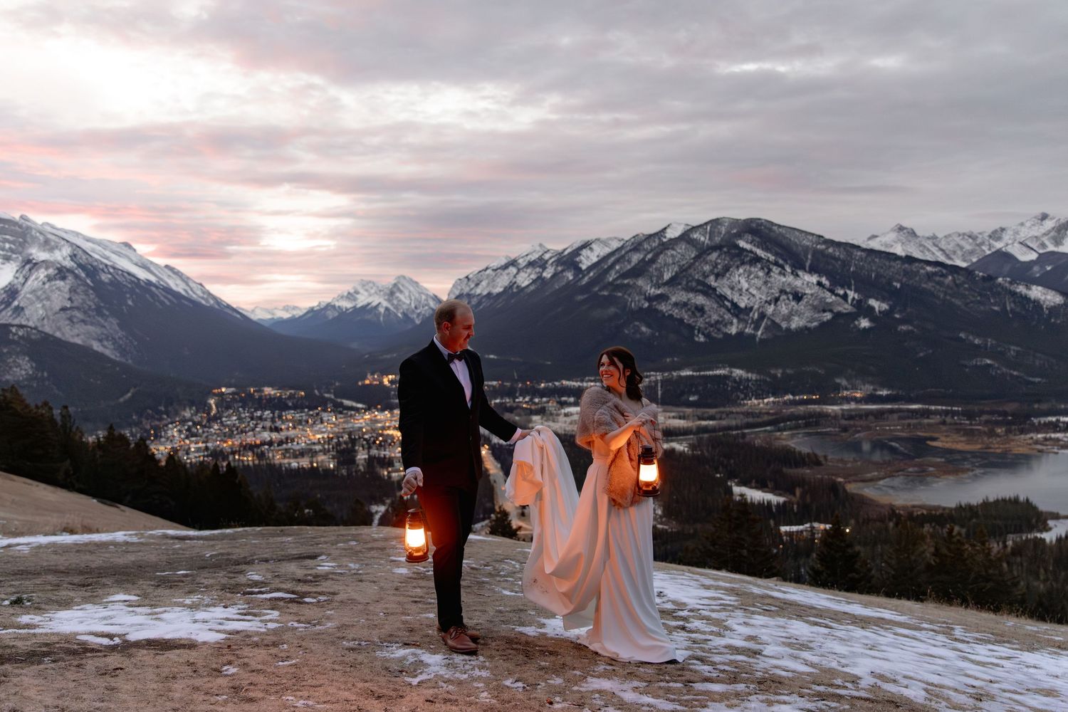 A couple stands on a snowy mountain overlook at sunset with the Canadian Rockies and Banff townsite visible in the distance.
