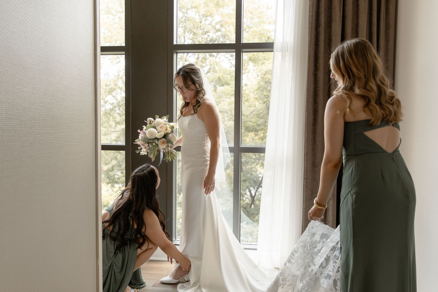 A bride in a white wedding dress stands by a window while getting ready for her wedding ceremony.