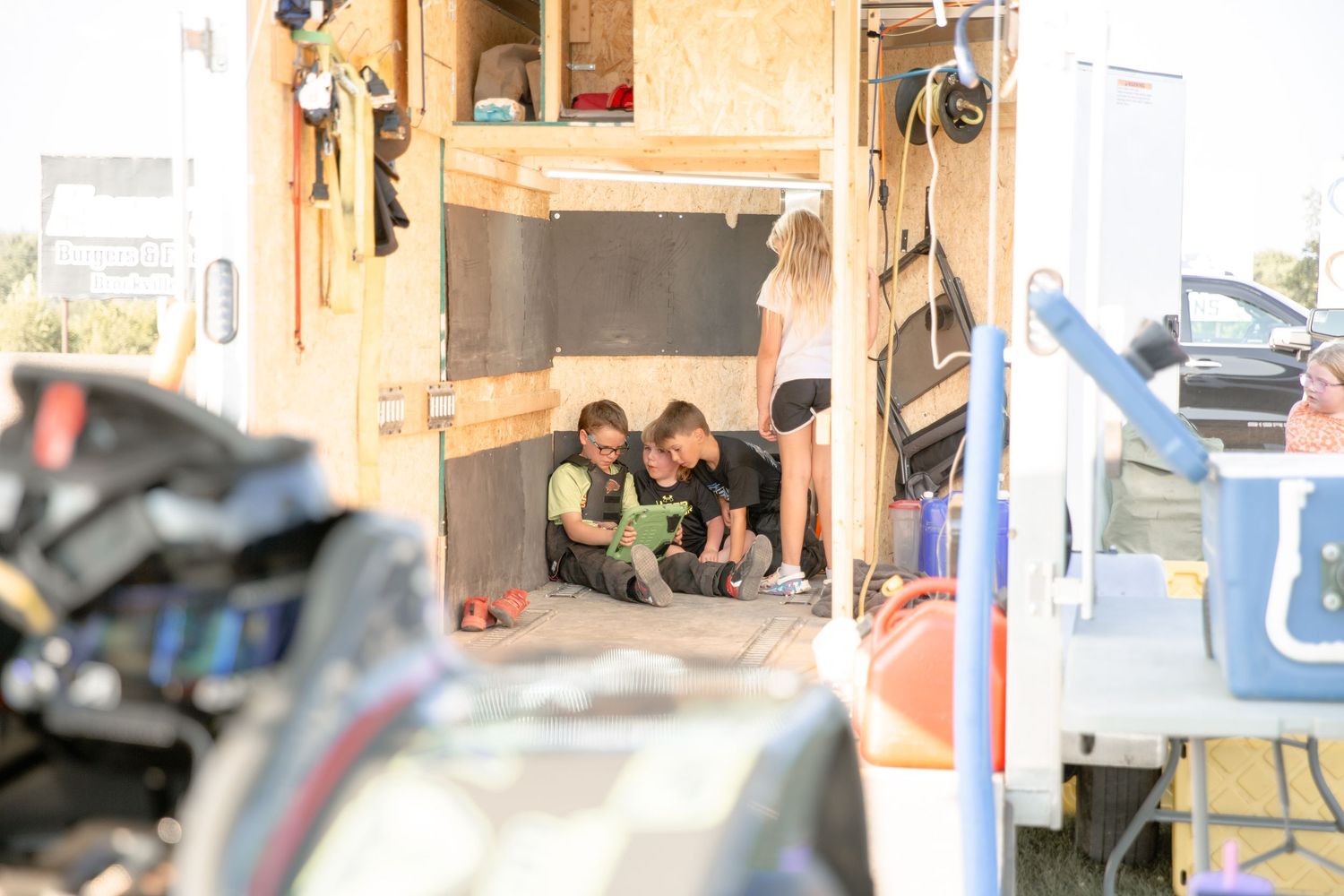 Children peek out from inside a cluttered storage space with tools and equipment visible in the foreground.