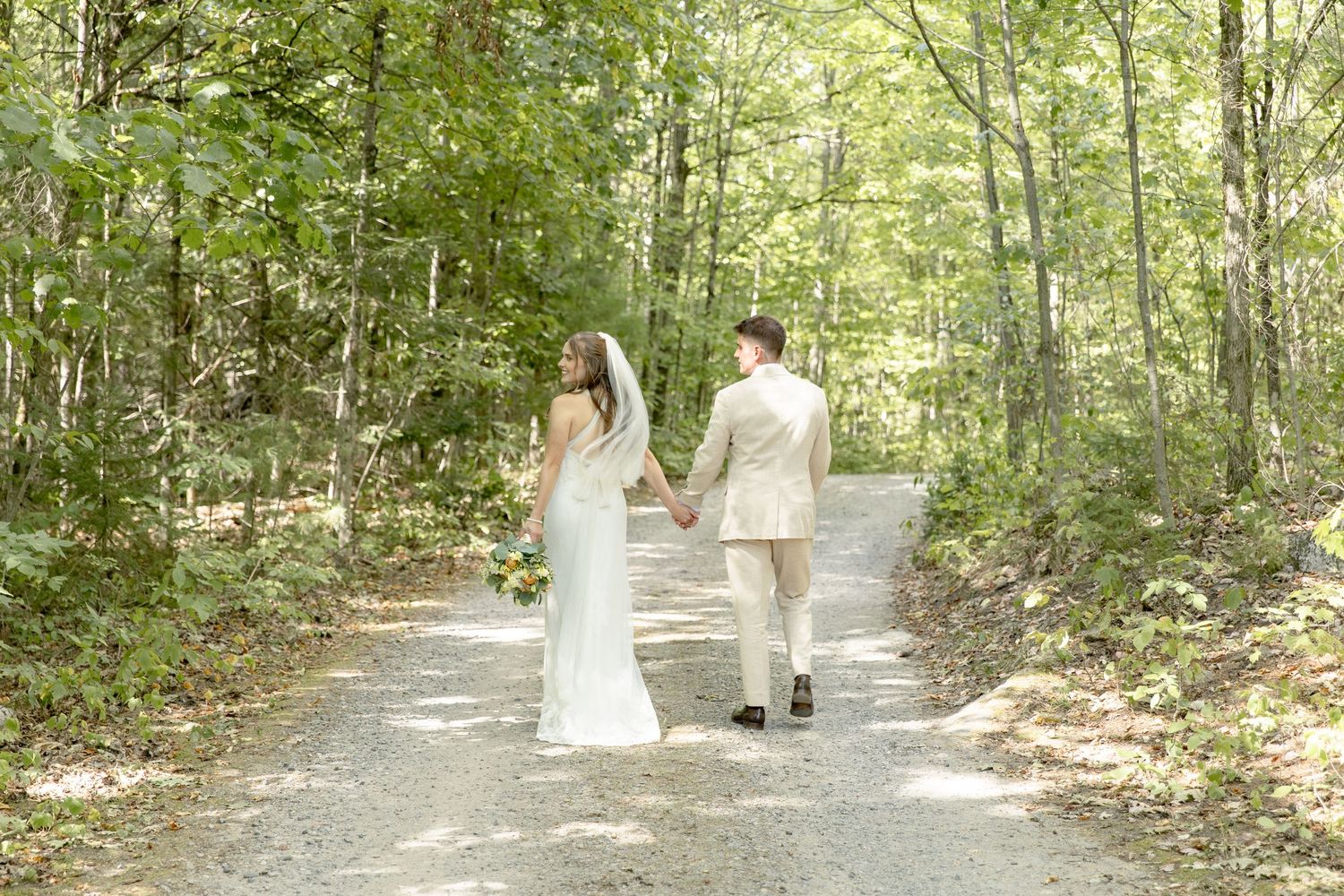 A couple walks hand in hand down a sunlit wooded path surrounded by lush green trees and foliage.