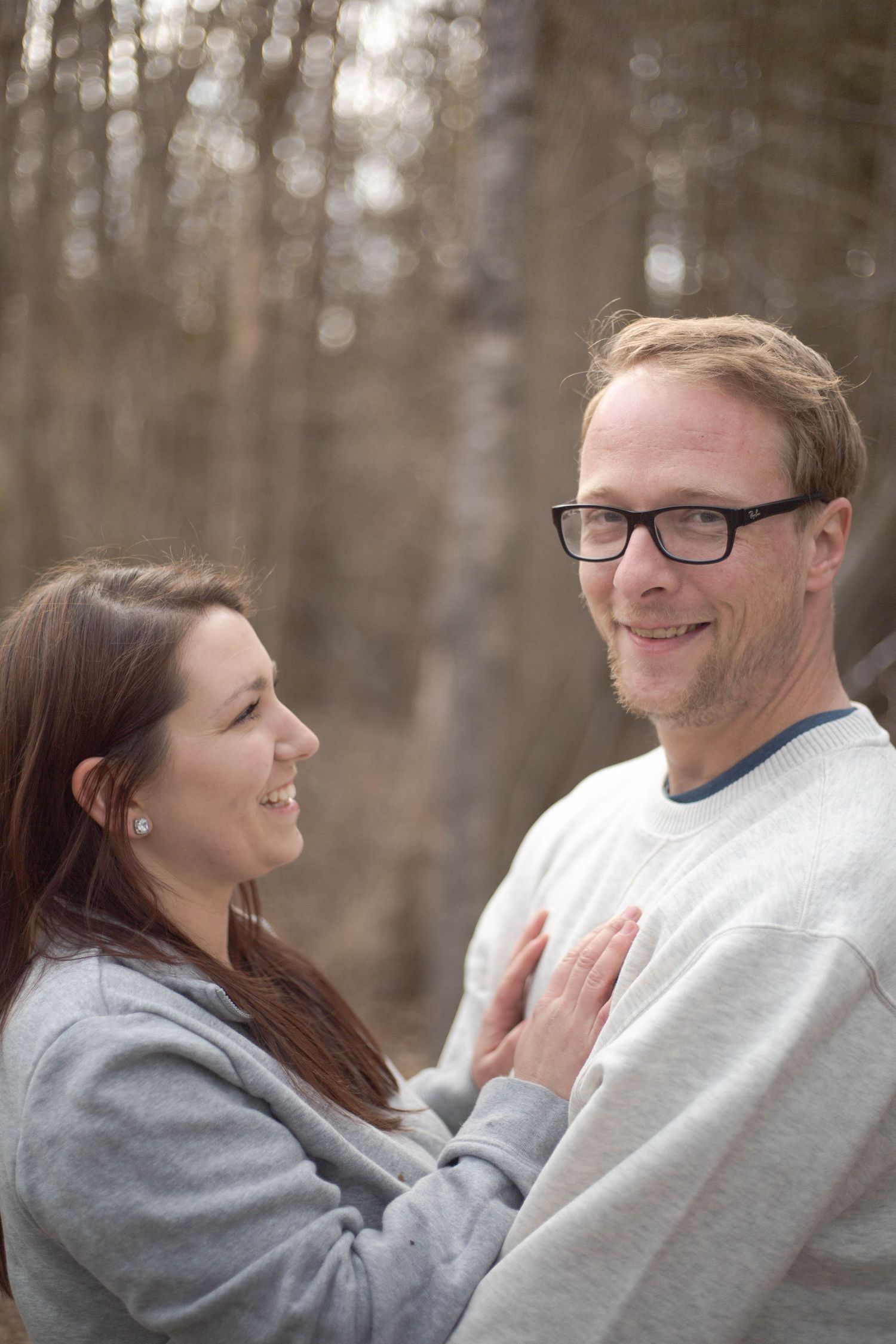 A couple shares an intimate moment outdoors in a wooded area, both wearing casual sweaters.