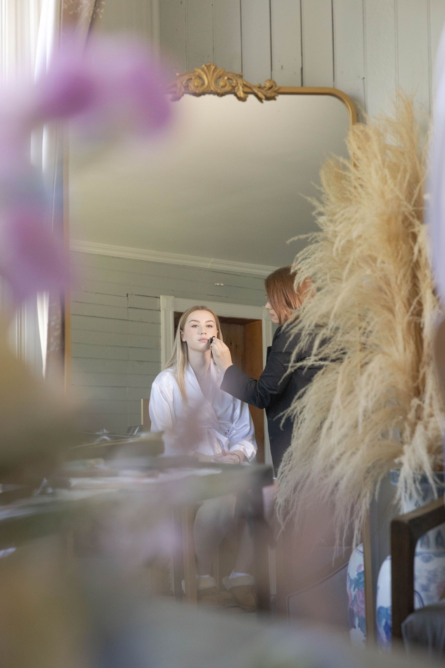 Reflection in an ornate mirror shows people getting ready, with pampas grass decor and soft purple flowers in foreground.