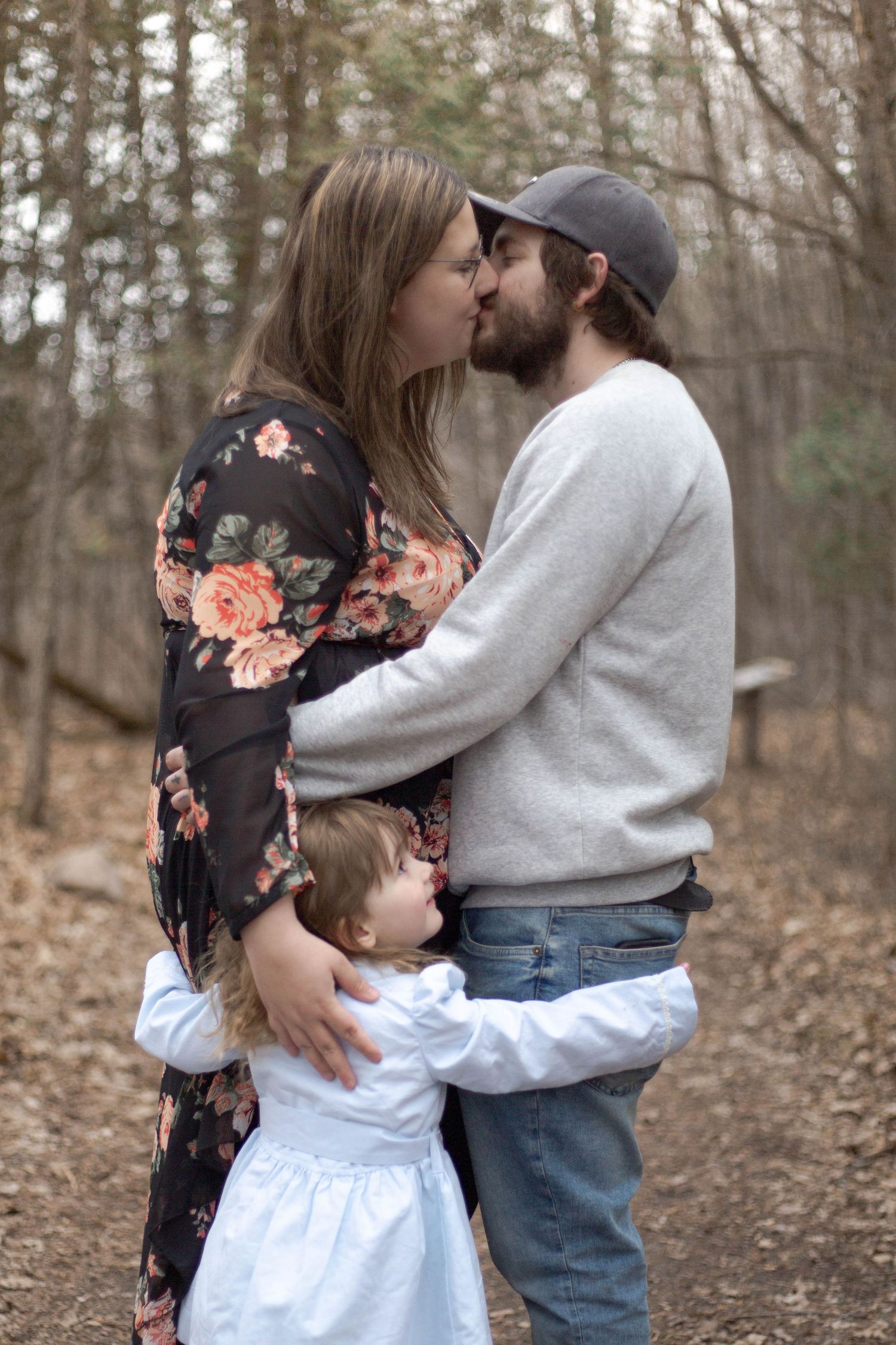 A couple shares a tender kiss in the woods while embracing a small child in a white dress.