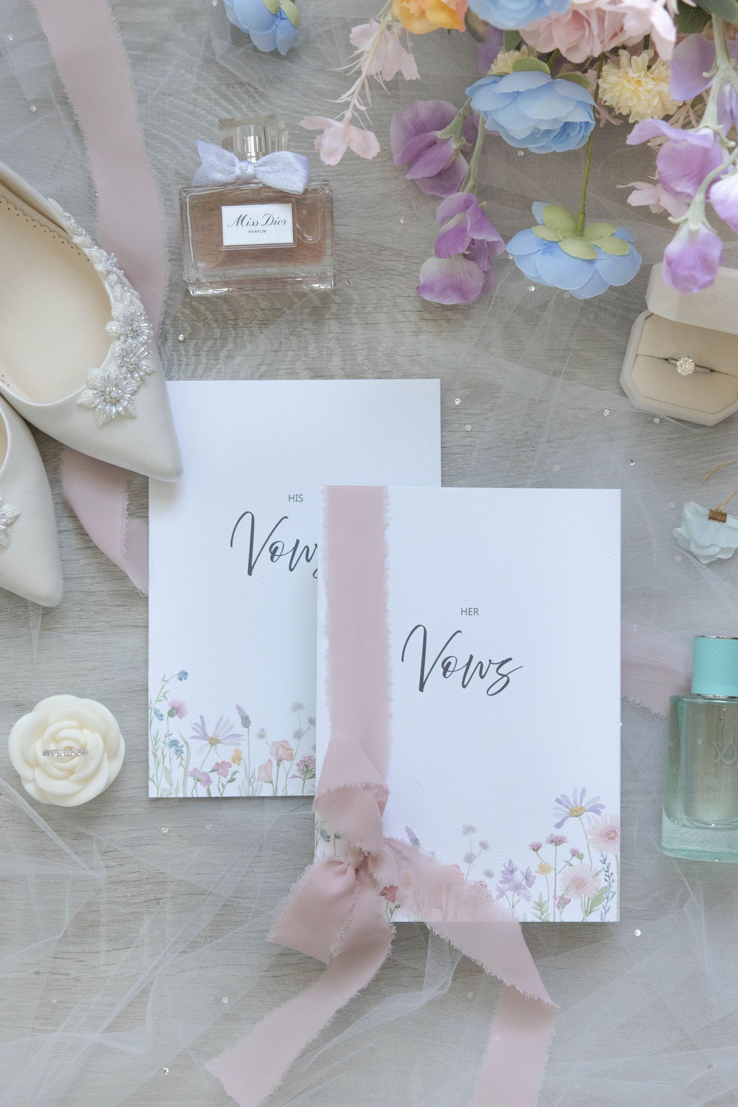 Wedding vow books with floral design tied with pink ribbon alongside perfume bottles and scattered flower petals.