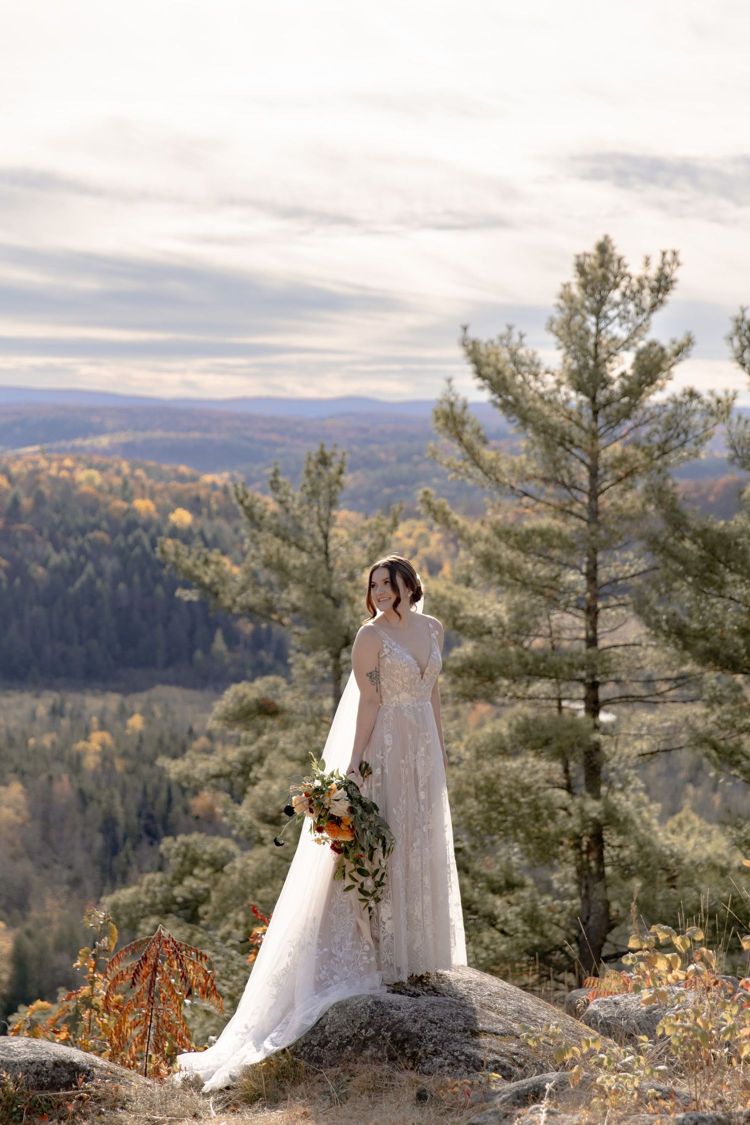 Two people in wedding attire stand on a rocky overlook with sweeping mountain views and autumn foliage at sunset.