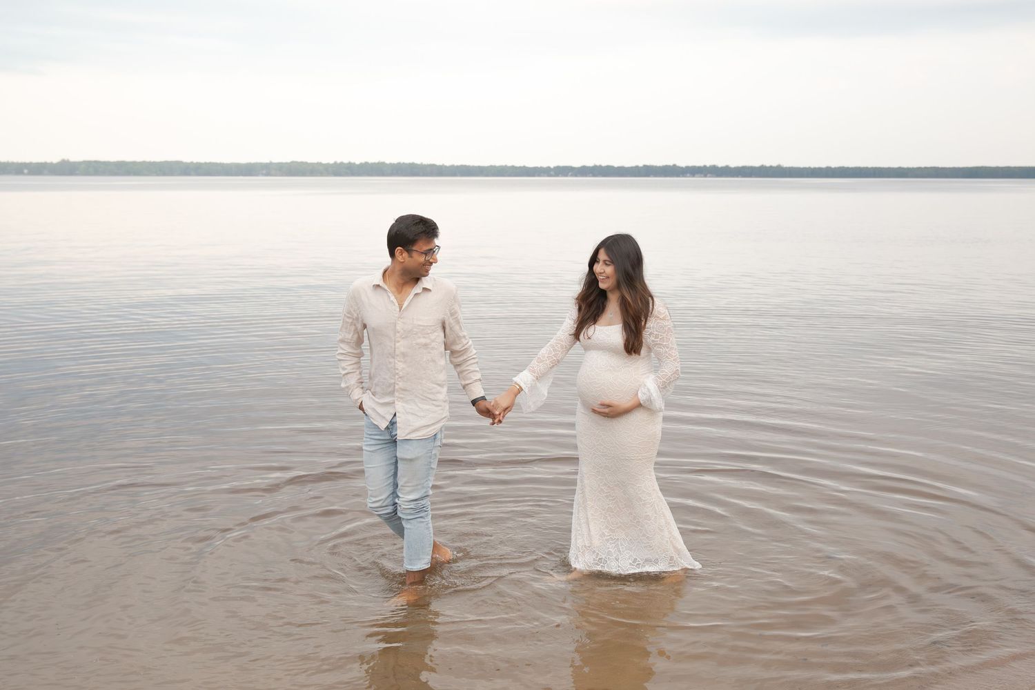 A couple stands in shallow ocean water at the beach, holding hands and wearing white clothing.