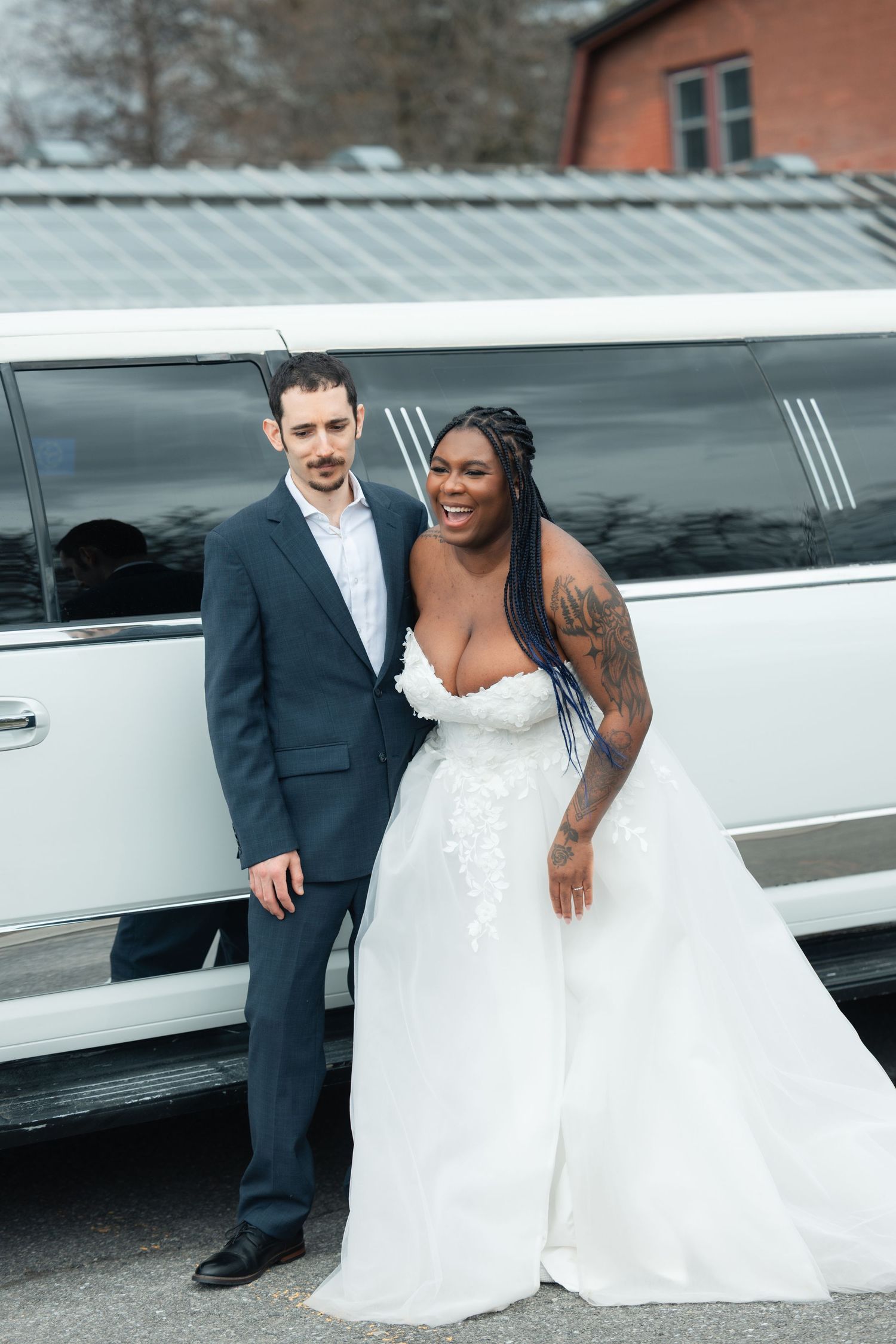 Interracial couple in wedding attire posing in front of a white limousine, bride in strapless gown with tattoos.