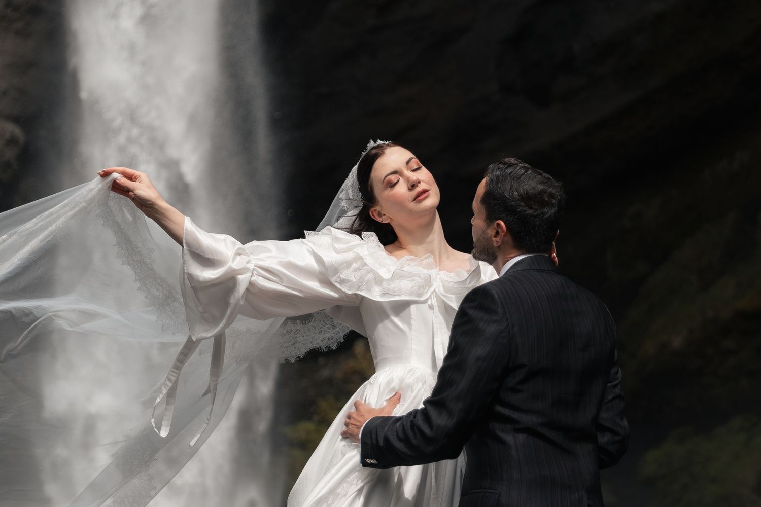 Bride in flowing white wedding gown and veil dancing near a waterfall with groom in dark suit holding her waist.