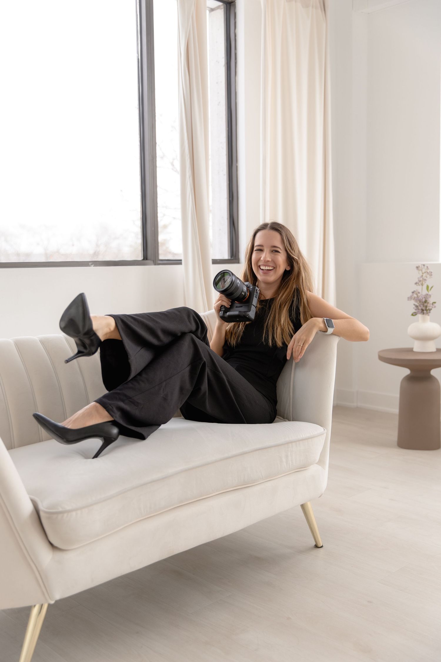 A person in a black dress lounges playfully on a modern white sofa in a bright minimalist room.