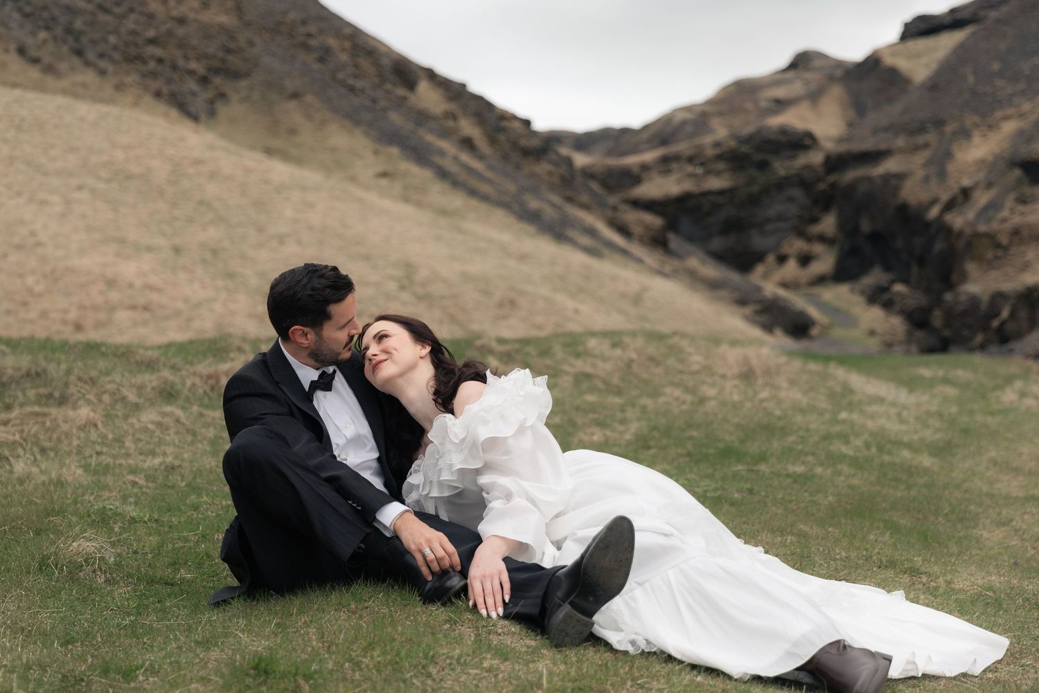 Bride in white gown and groom in tuxedo sitting on green hillside in Iceland during outdoor wedding photoshoot.