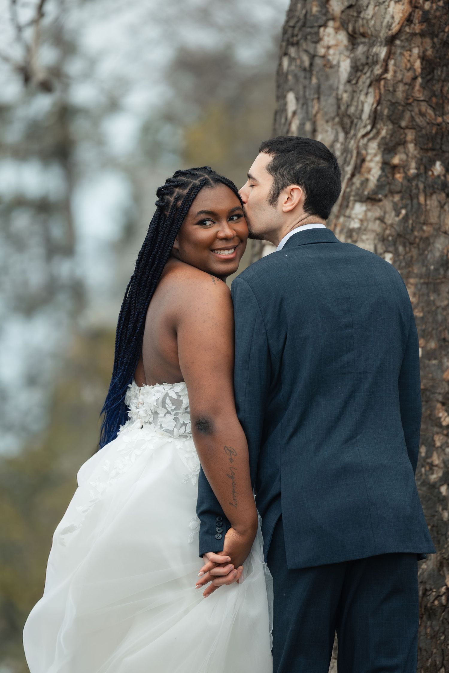 Interracial couple in wedding attire pose by a tree, bride in white gown smiles as groom kisses her cheek.