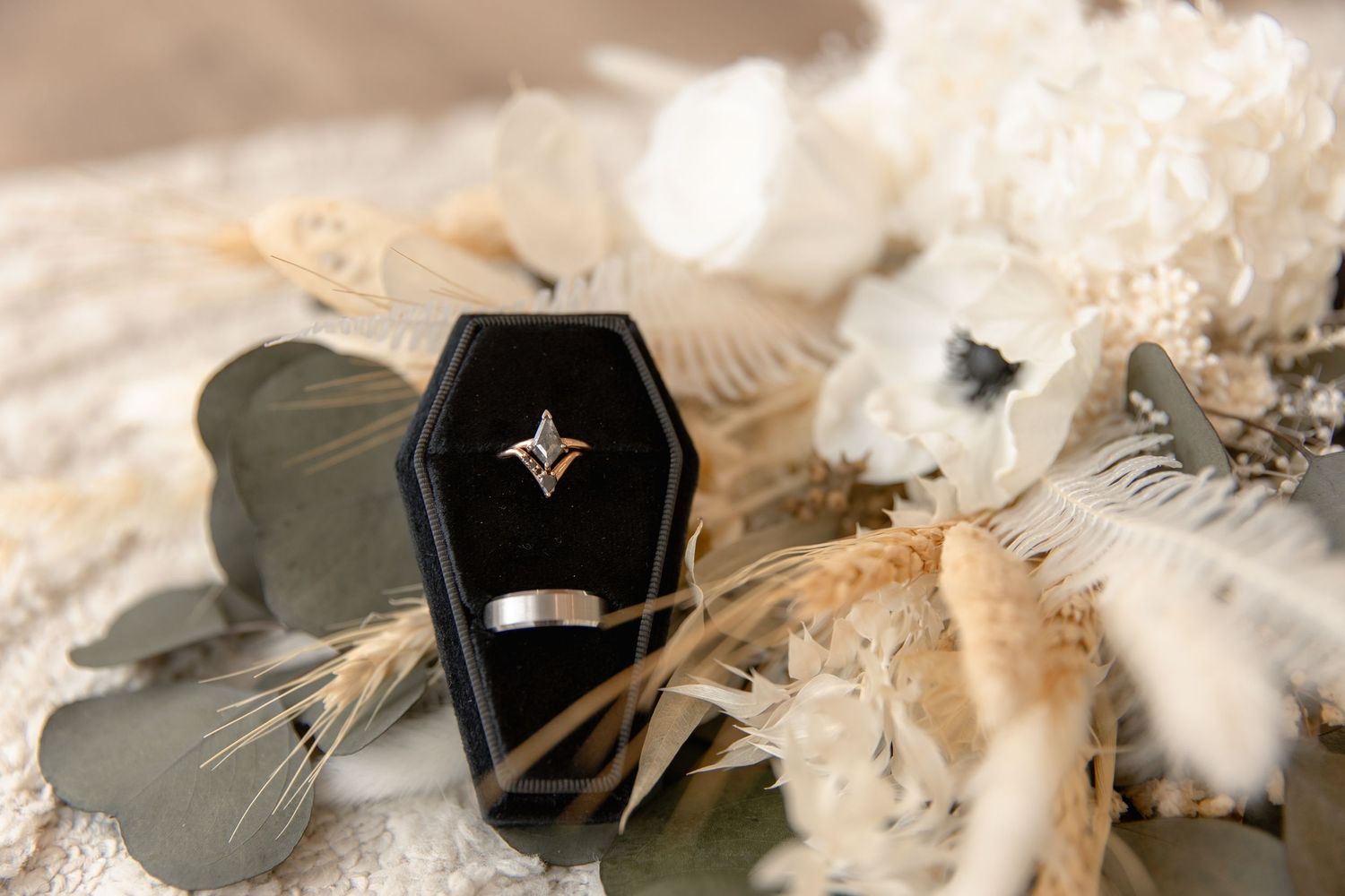 Black coffin-shaped ring box with engagement ring and dried flowers on beige fabric.