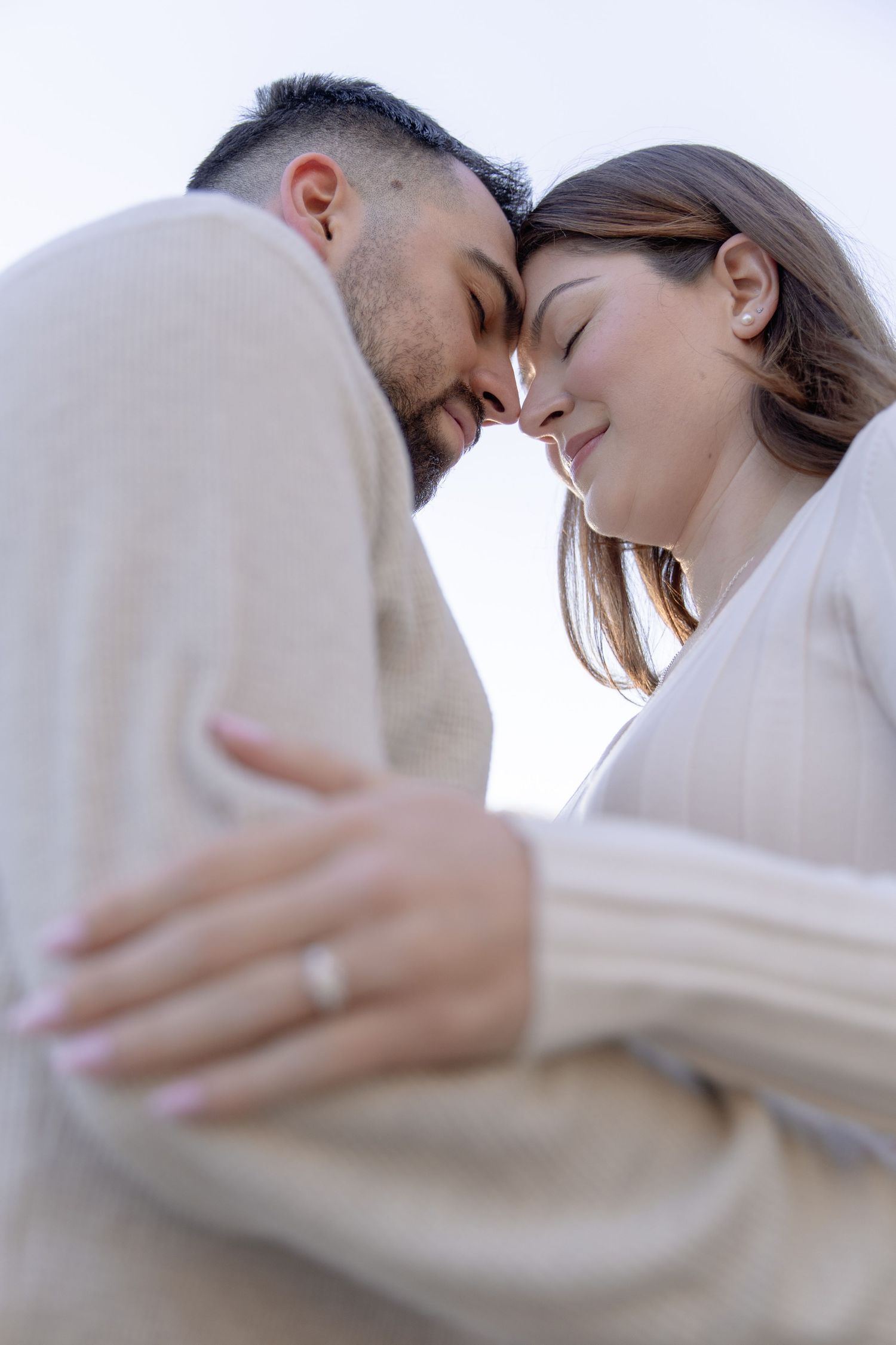 Romantic couple in beige sweaters share an intimate forehead touch against bright sky background.