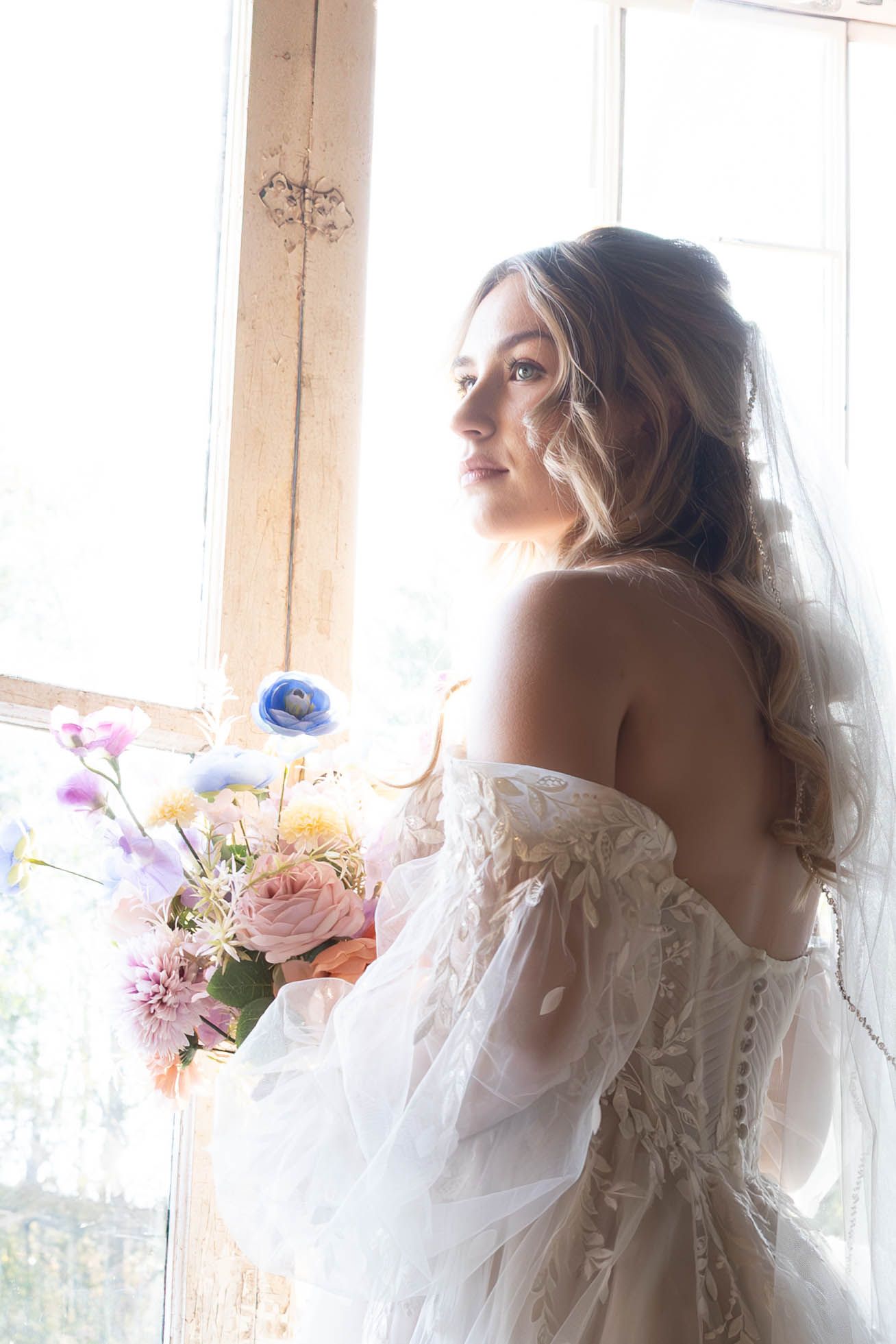 Bride in lace off-shoulder wedding gown holding pastel bouquet poses by window in dreamy natural light.