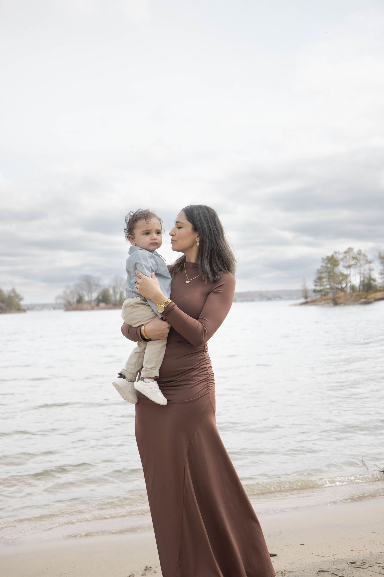 Someone wearing a long brown dress holds a young child while standing on a beach shoreline on an overcast day.