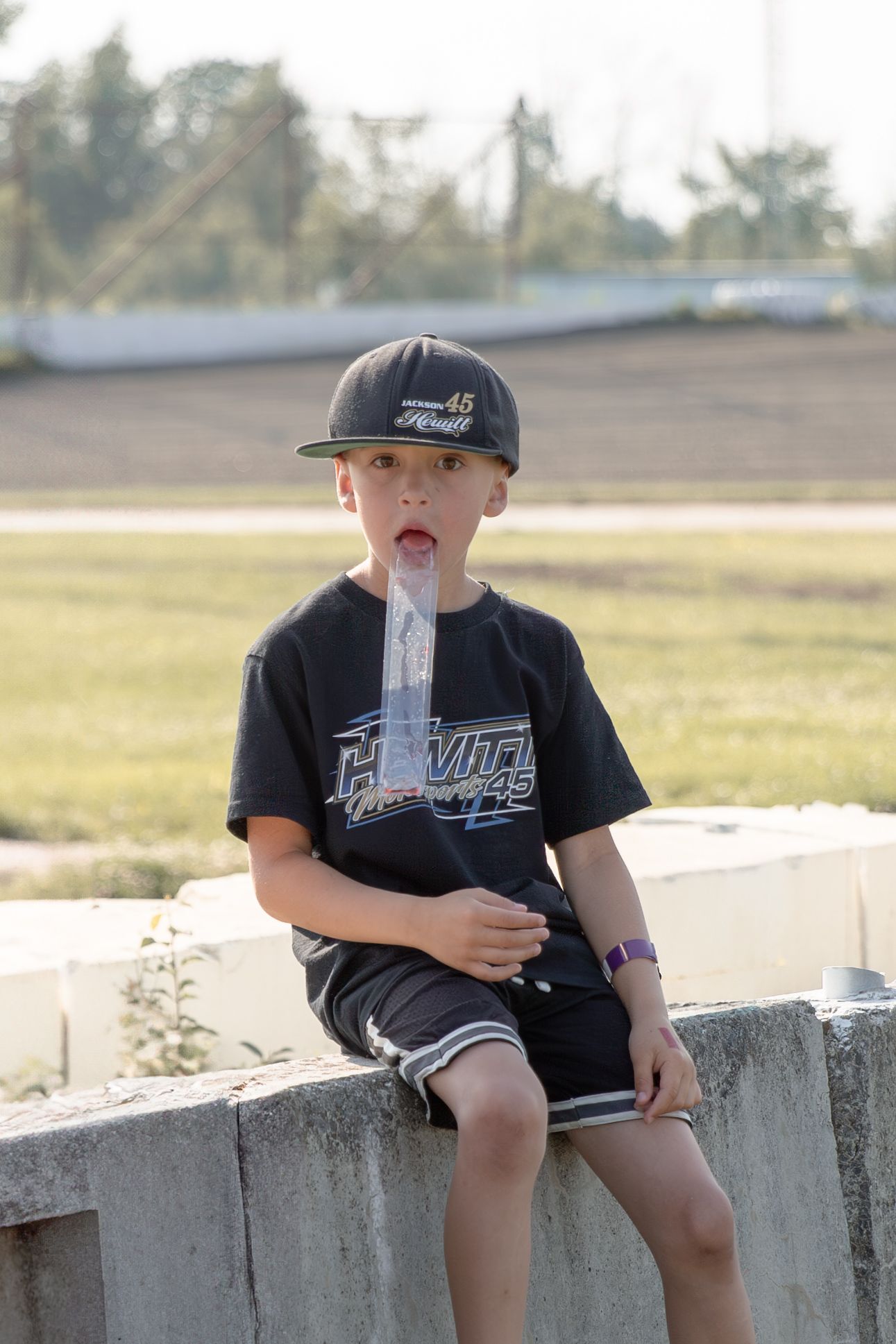 Child in black t-shirt and baseball cap sits on concrete wall while drinking from a water bottle on sunny day.