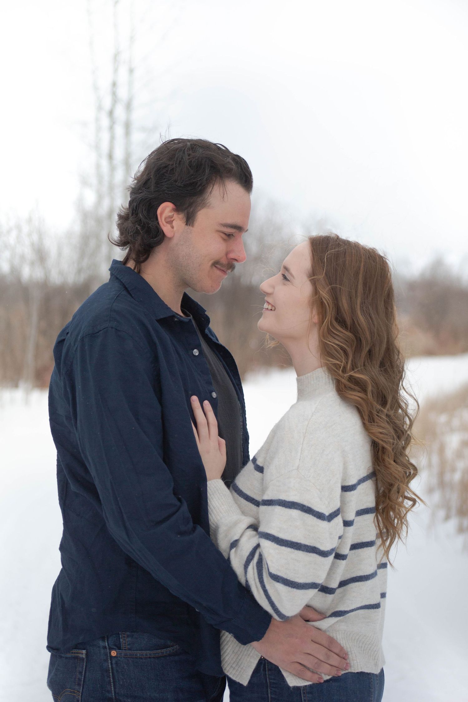 A romantic winter engagement portrait of a couple in casual attire sharing an intimate moment in a snowy field.