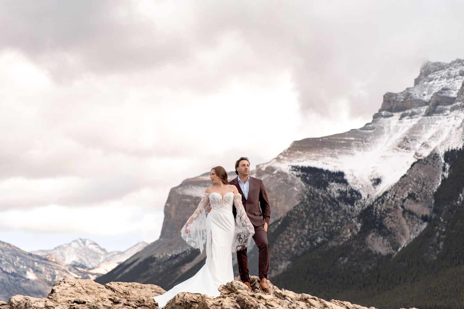 A couple in wedding attire stands on a rocky mountain peak with snow-capped Canadian Rockies mountains in the background.
