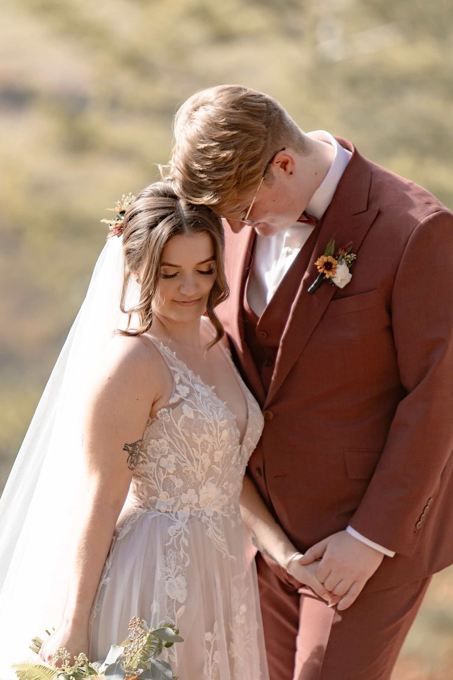 A romantic wedding couple embraces in a desert landscape wearing burgundy suit and lace gown during golden hour.