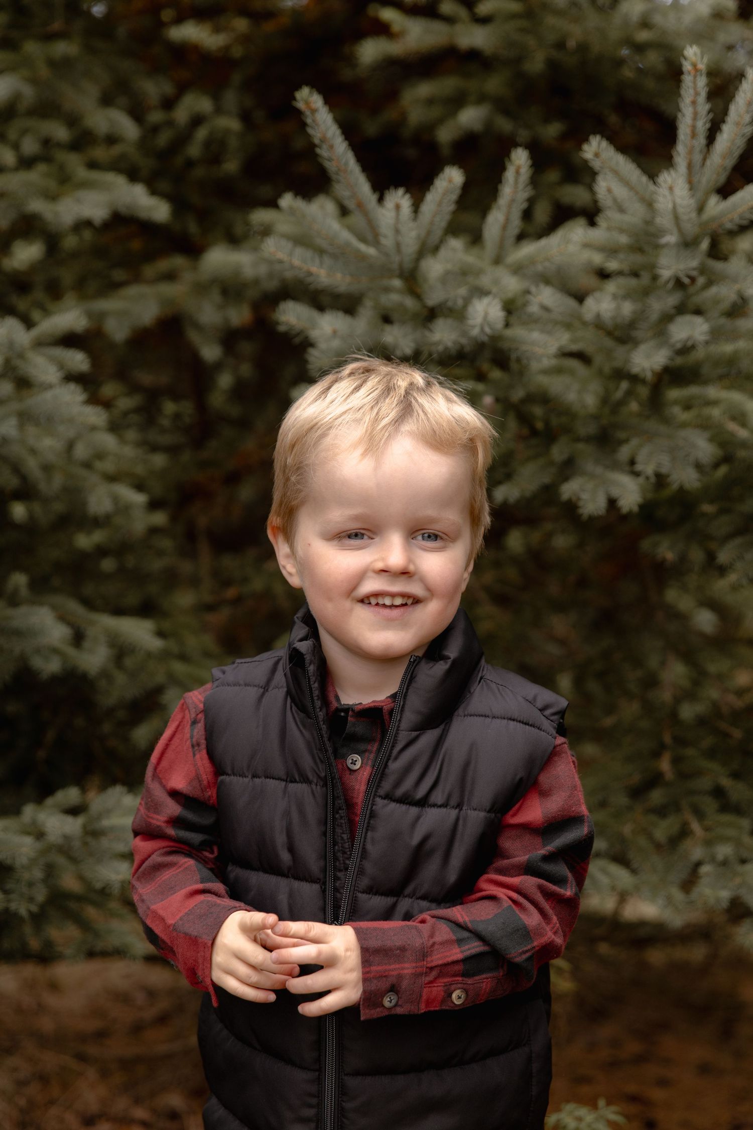 A child wearing a black vest and red plaid shirt smiles warmly against an evergreen tree background.