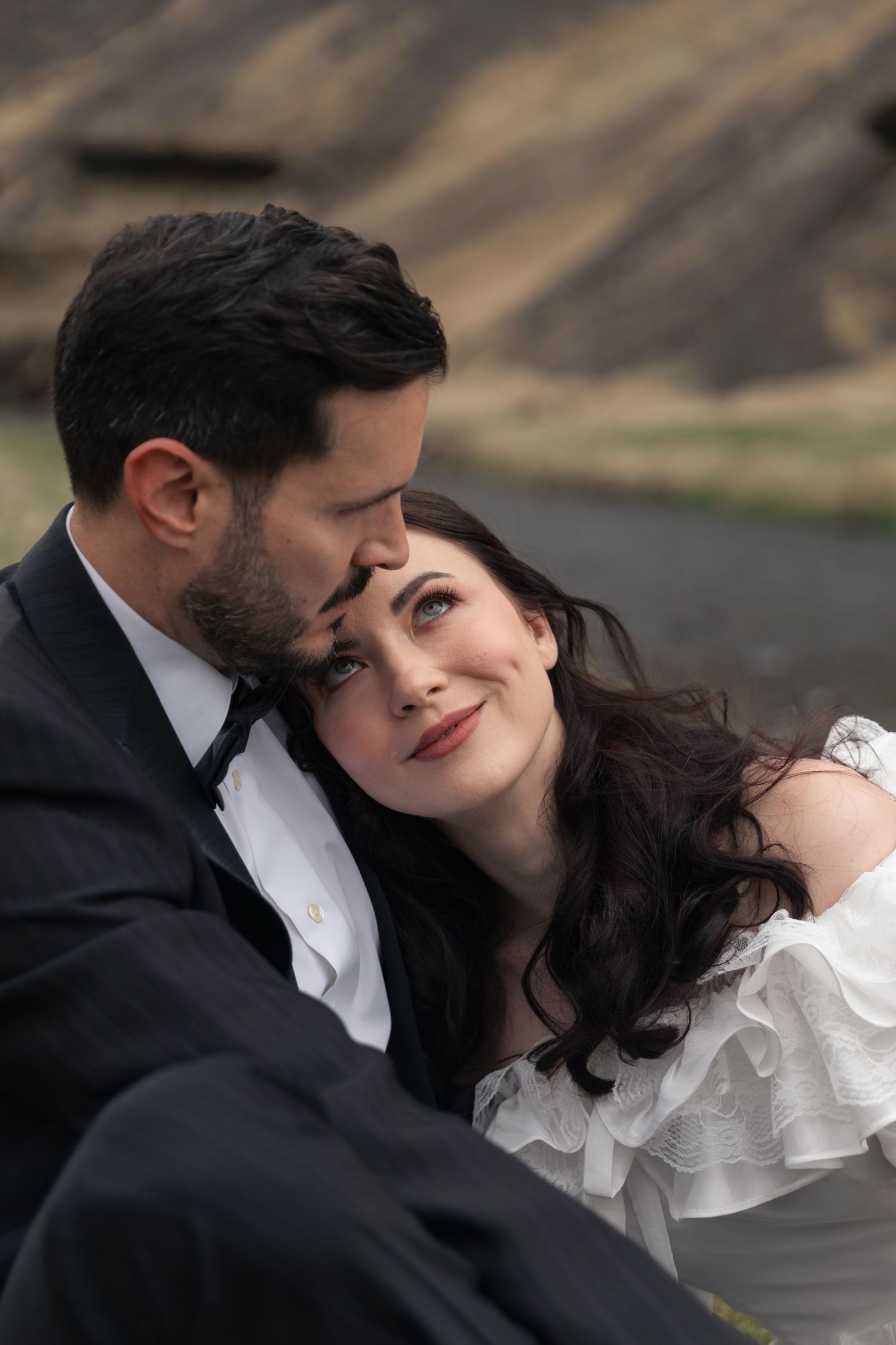 Romantic couple in wedding attire embracing outdoors, bride in white ruffled dress smiling up at groom in dark suit.