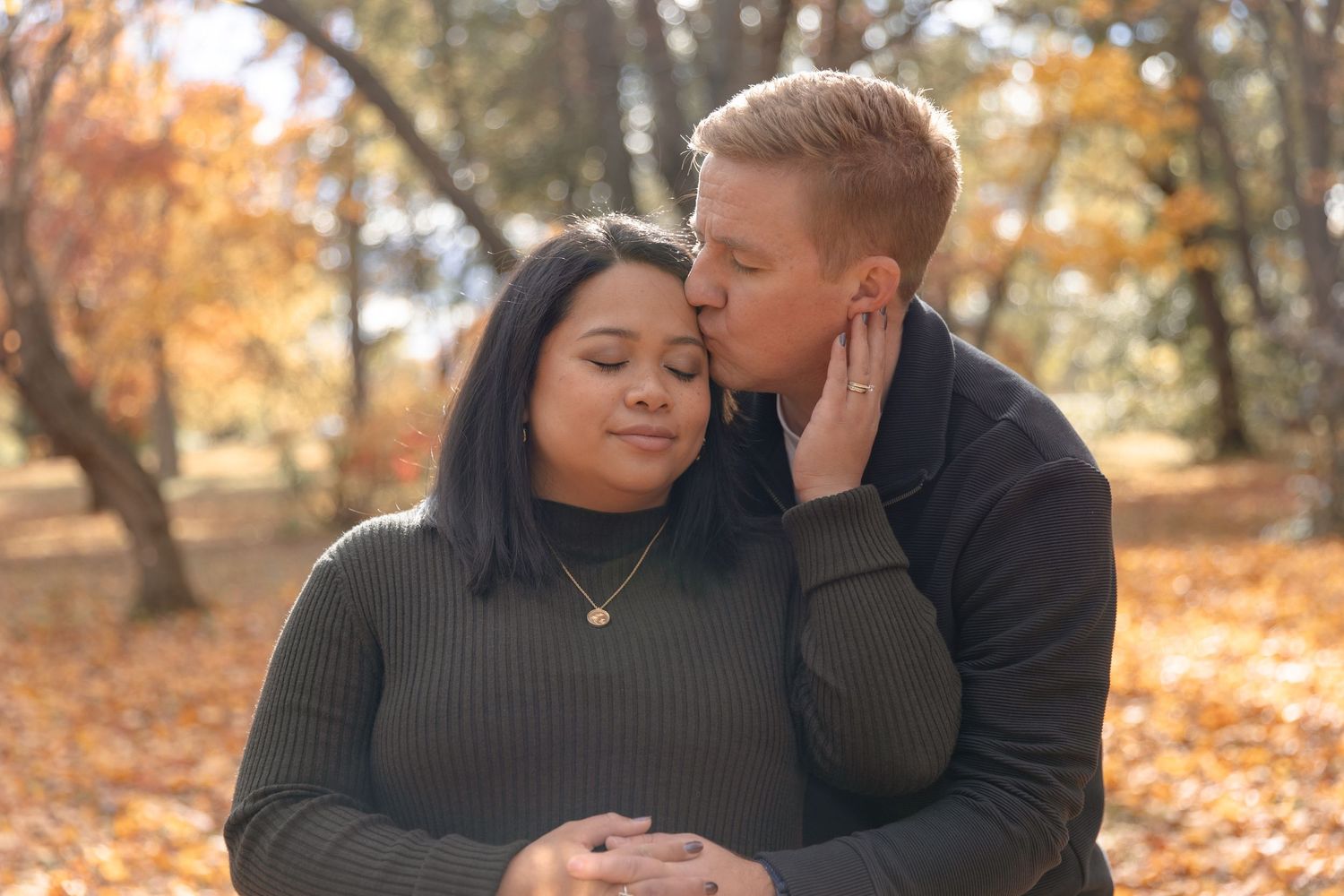 A couple shares an intimate moment outdoors surrounded by autumn foliage and fallen leaves in a park setting.