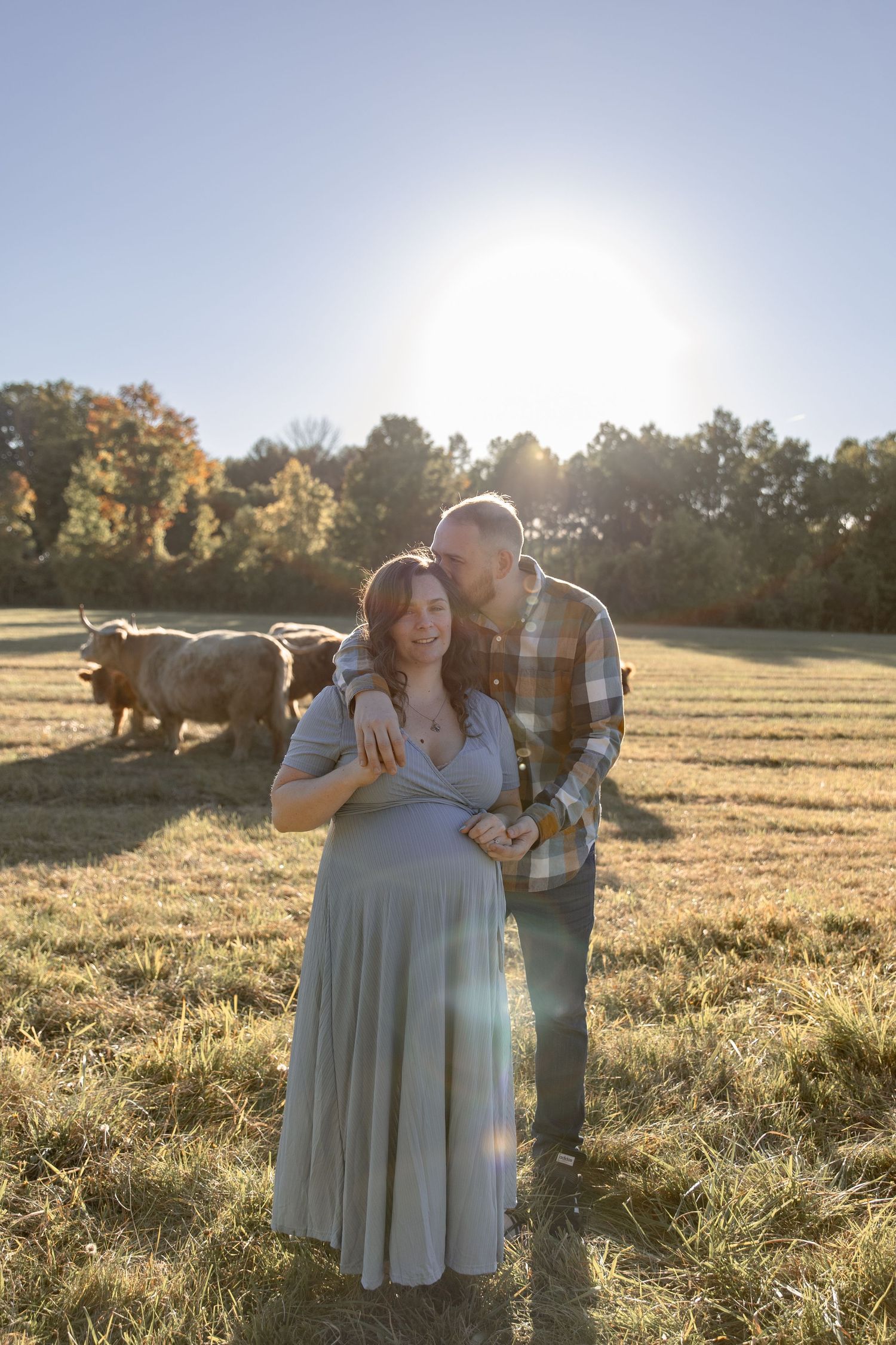 A couple embraces in a sunlit field at golden hour as sunlight streams through autumn trees in the background.
