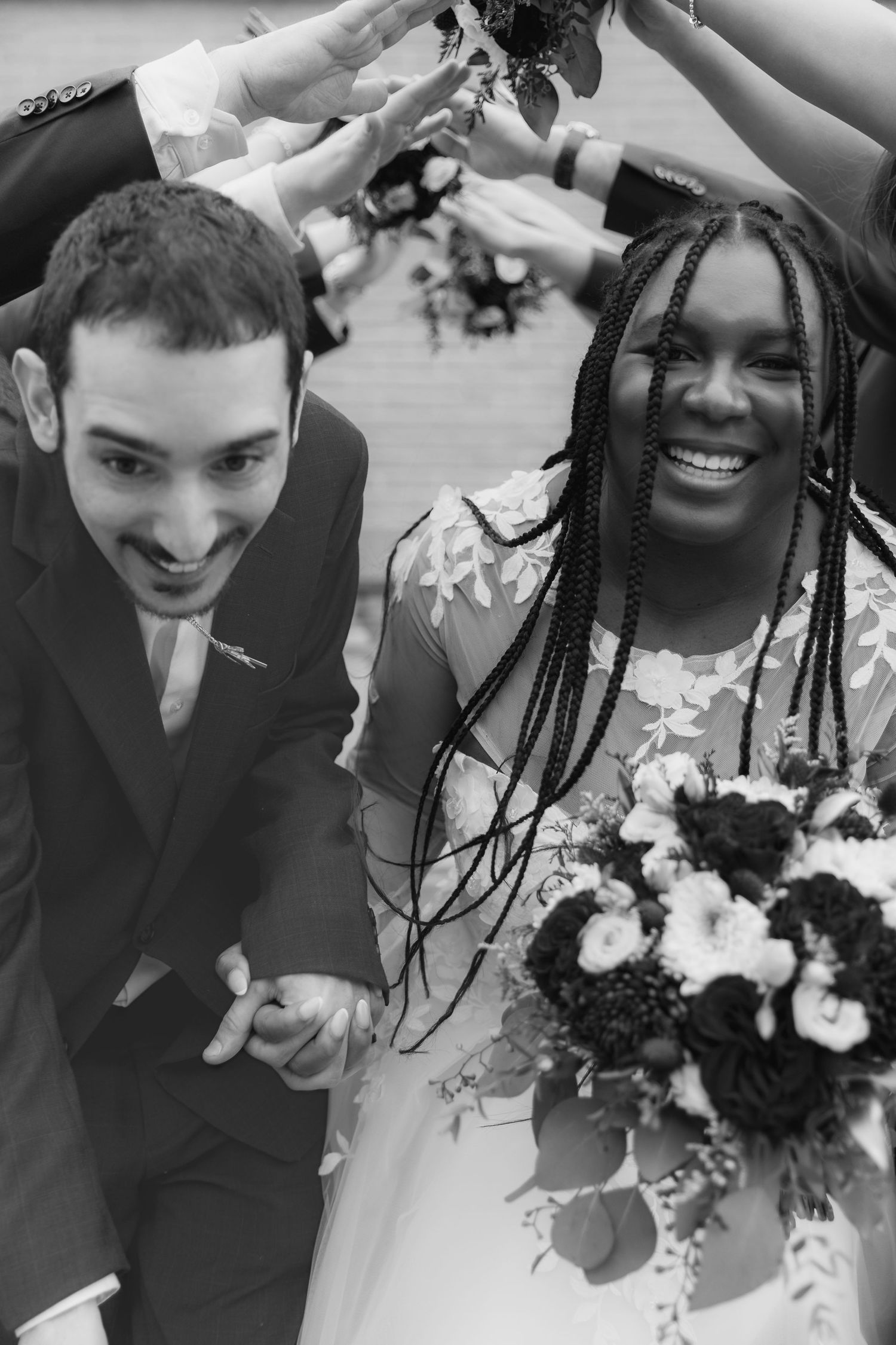 Black and white photo of a joyful interracial couple walking under an arch at their wedding ceremony.