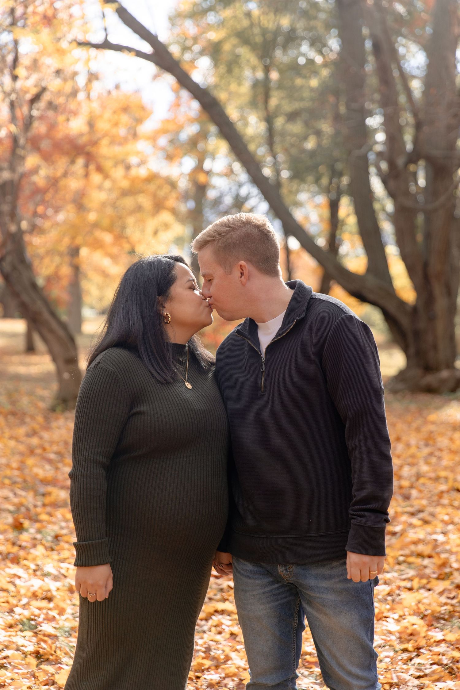 A romantic couple in black sweaters share tender moments among fallen autumn leaves in a park setting.