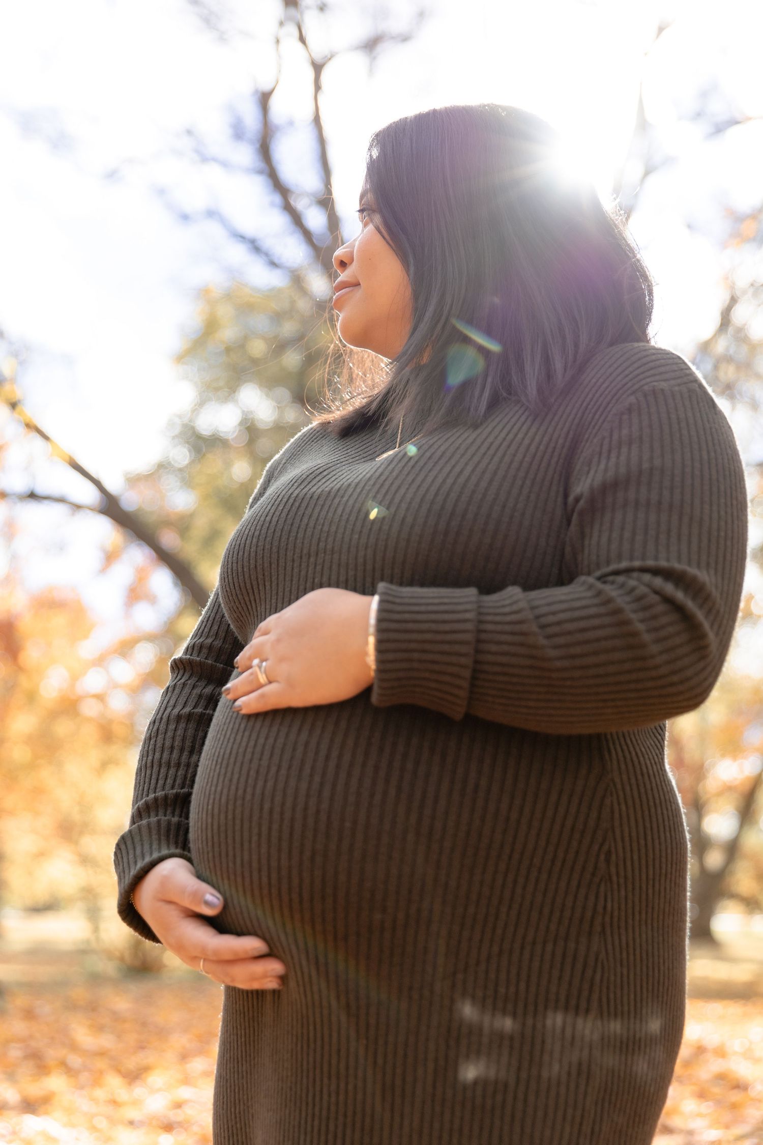 Person in black sweater cradling pregnant belly during autumn maternity photoshoot in park setting.