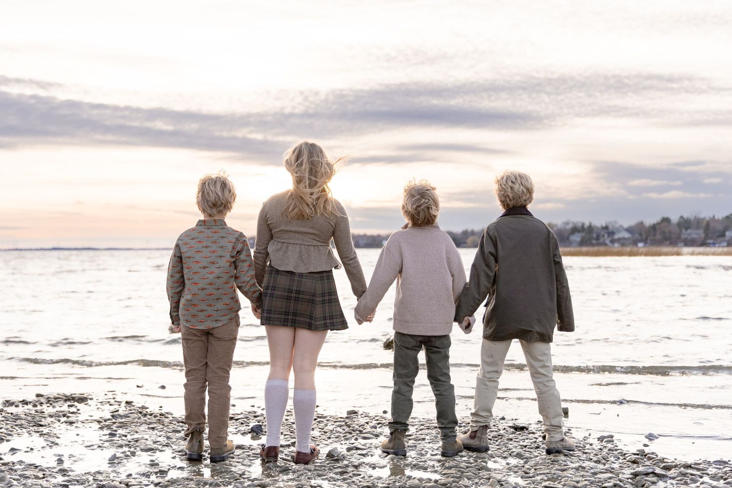 Four people hold hands while standing together on a beach at sunset looking towards the ocean.