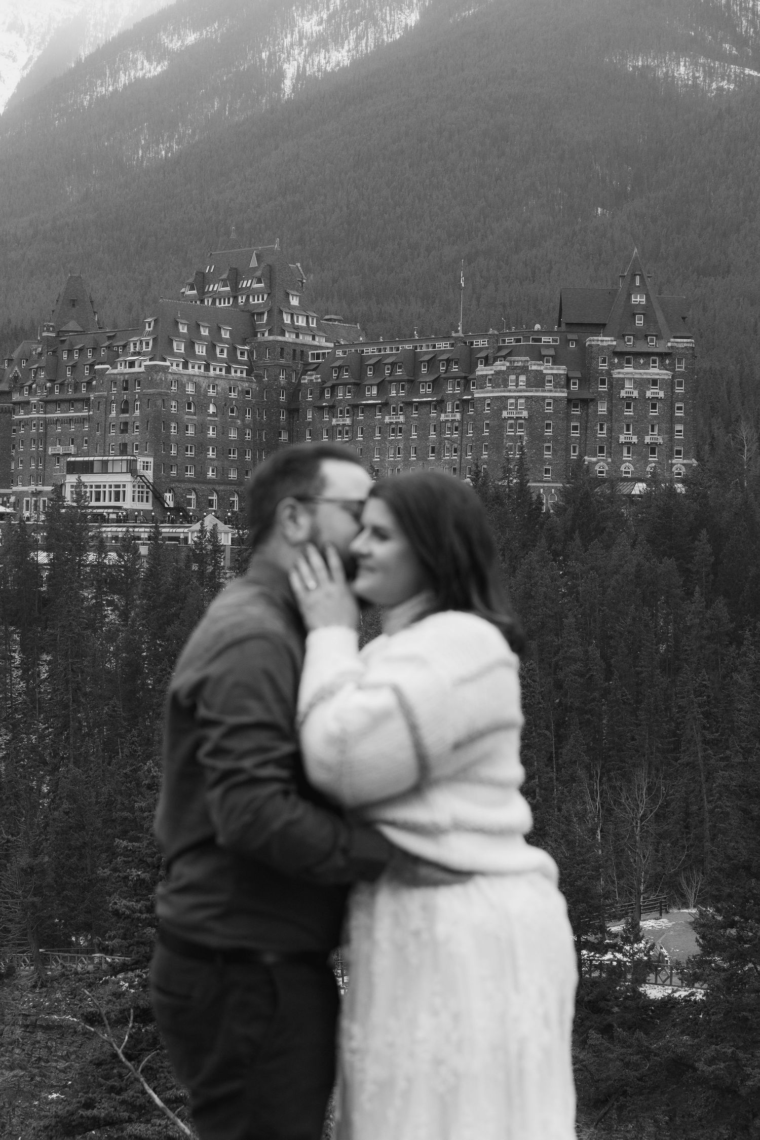 A romantic couple embraces in front of the historic Banff Springs Hotel with snow-capped mountains in the background.