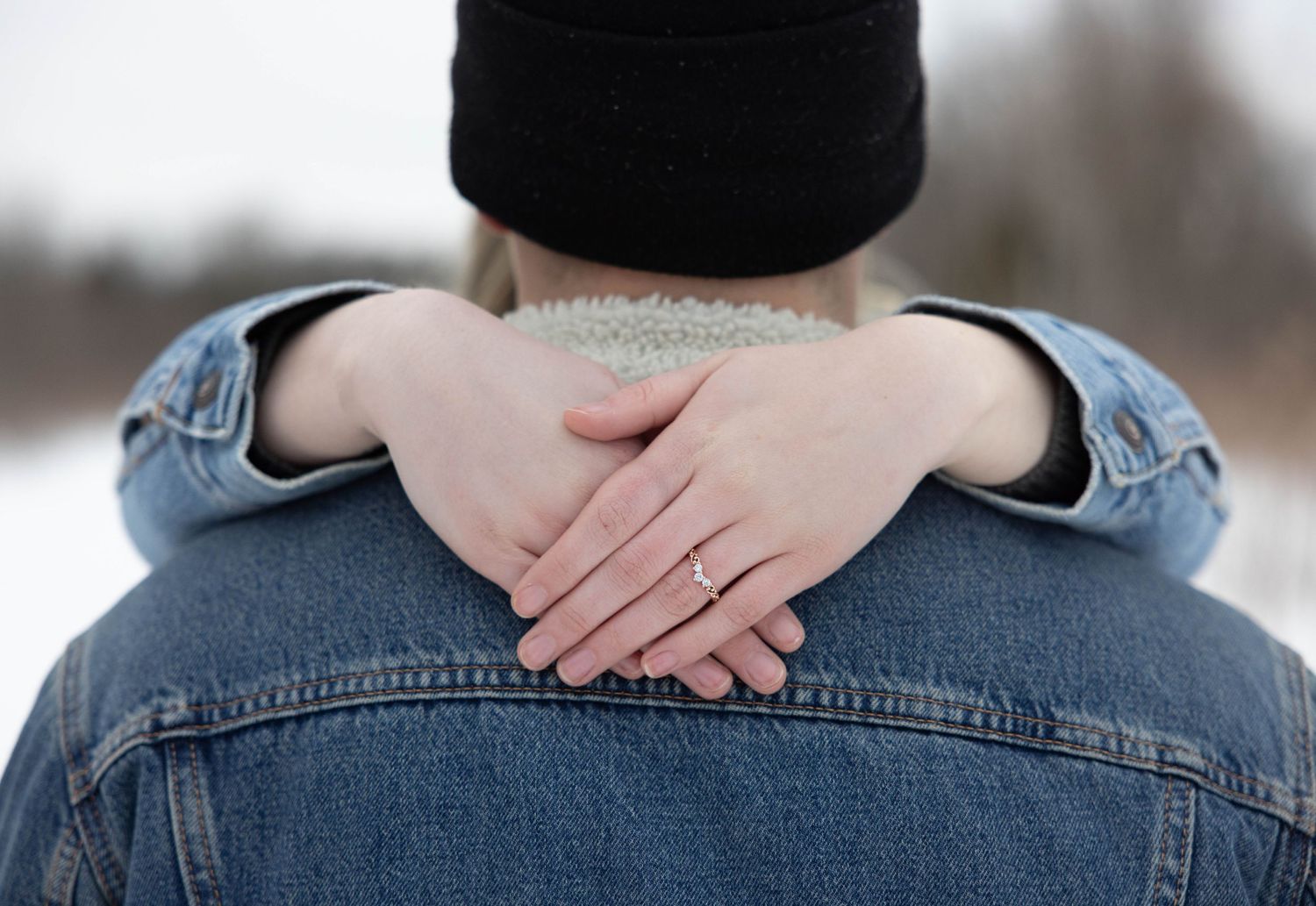 Close-up of hands with engagement ring resting on denim jacket behind fiancé's neck in an intimate winter portrait.