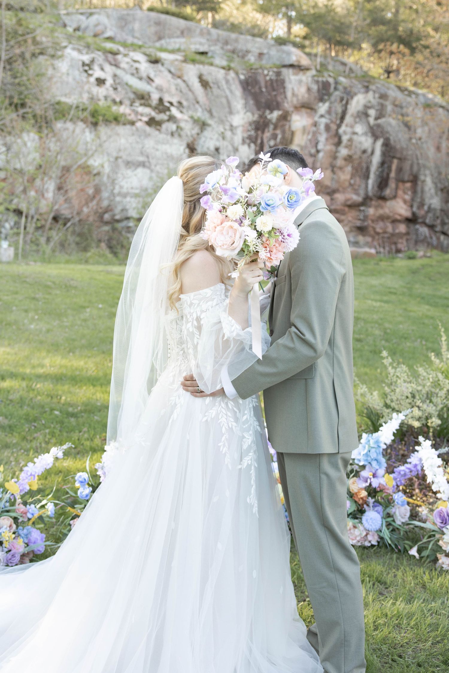 A romantic wedding couple embraces while holding a pastel bouquet against a rocky cliff backdrop with floral decorations.