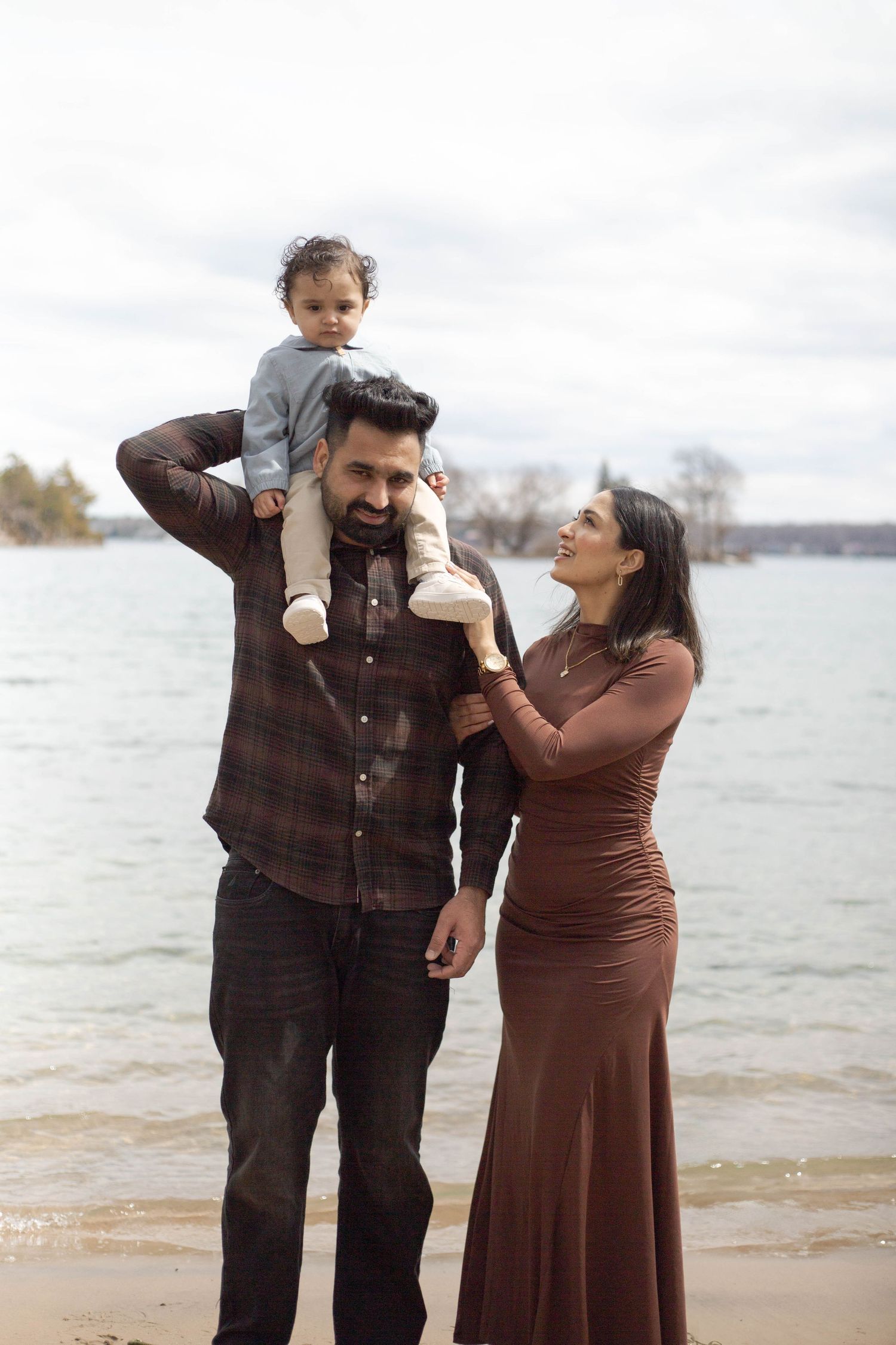 A family stands together at the beach on an autumn day, with a small child being carried on shoulders.
