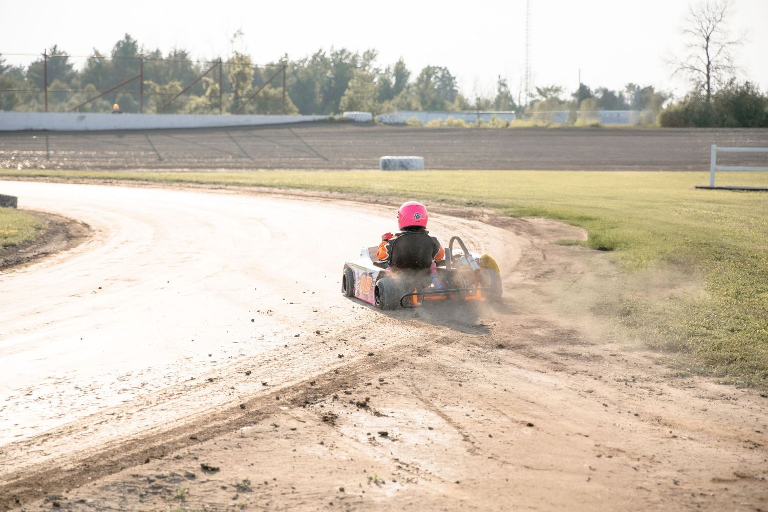 A go-kart kicks up dust while racing around a dirt track curve on a sunny day.