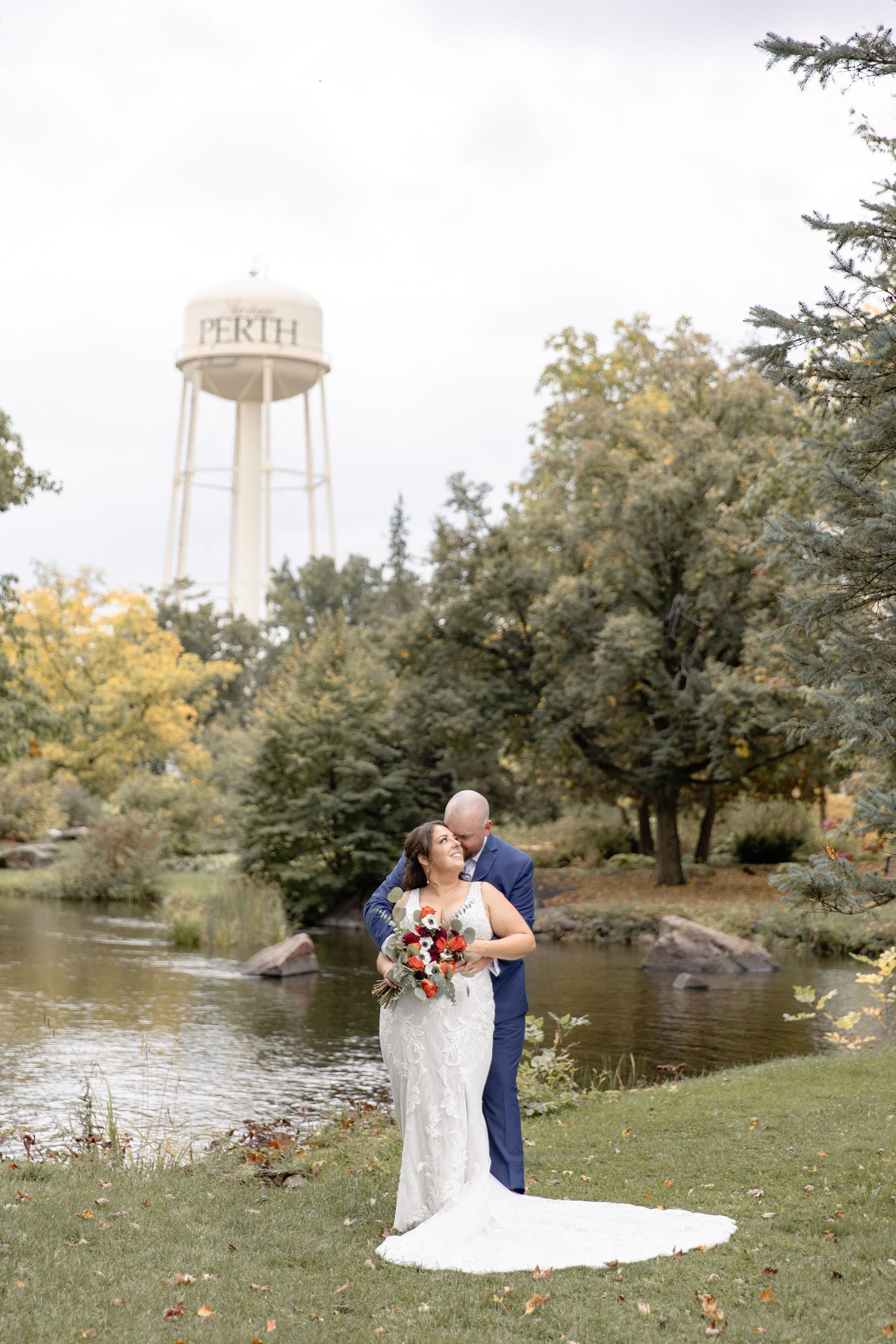 A scenic wedding photoshoot by a pond with a white water tower visible in the background through autumn trees.
