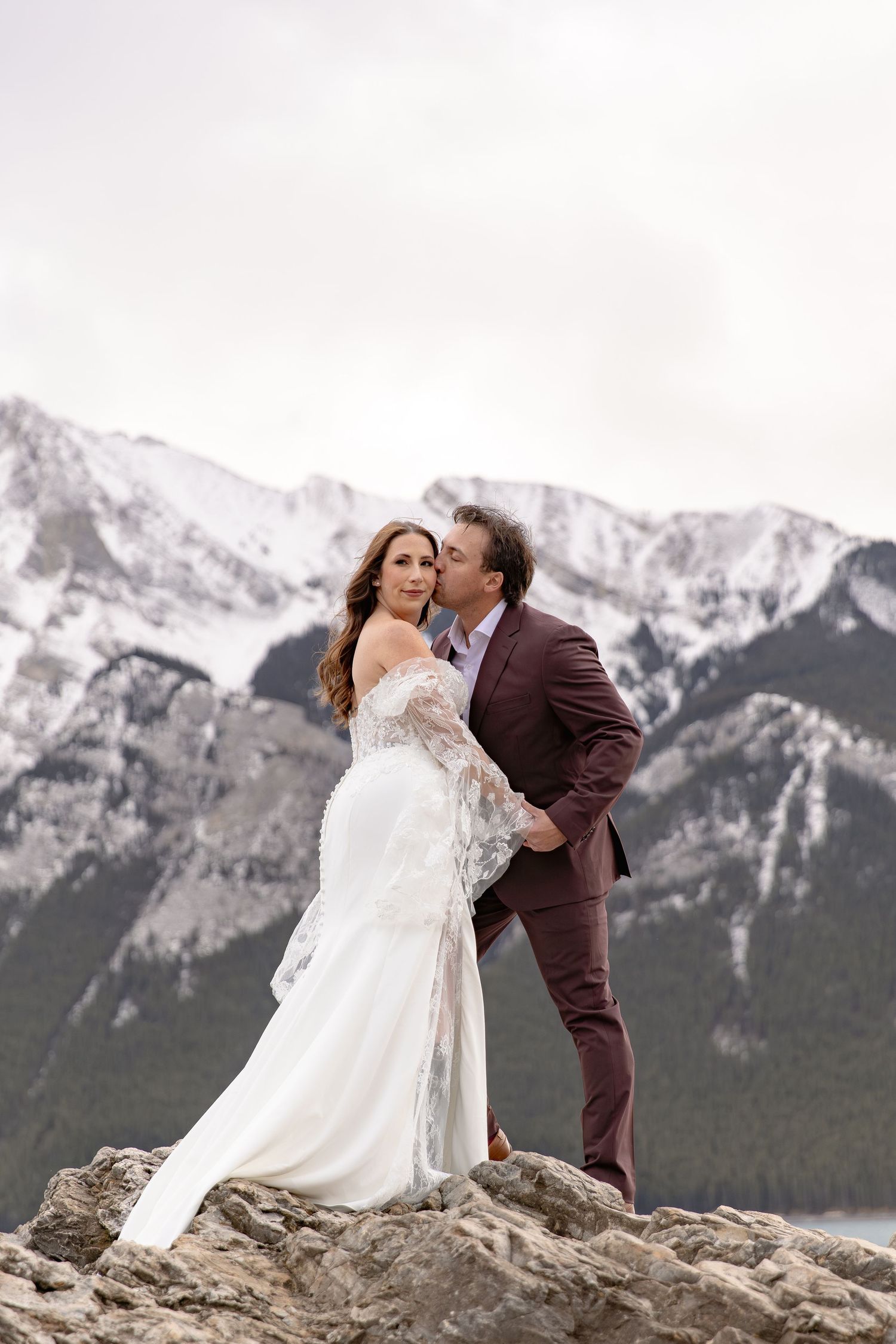 A romantic wedding couple shares an intimate moment on a rocky mountain overlook with snow-capped peaks in the background.