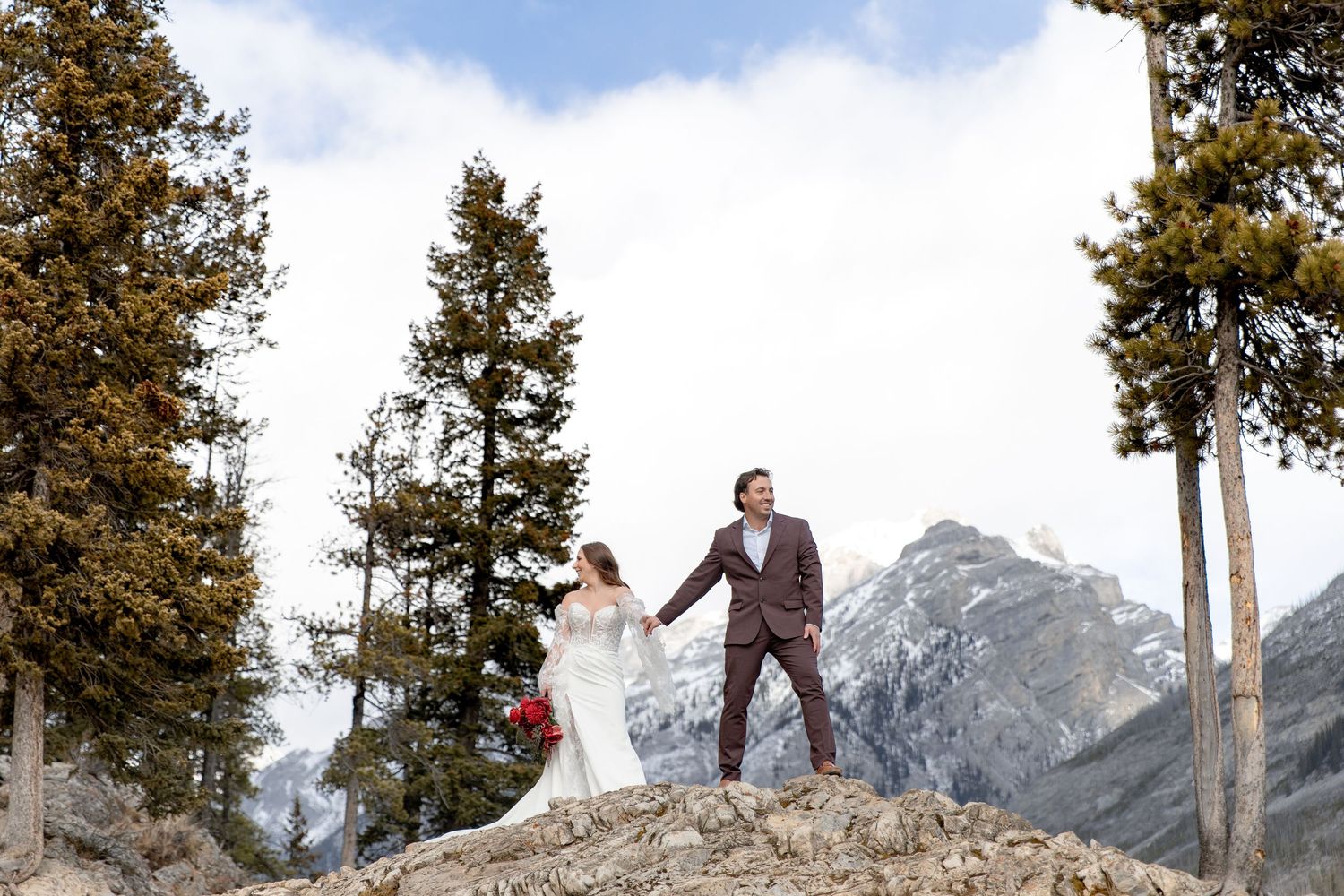 A wedding couple poses dramatically against a backdrop of snow-capped mountain peaks and evergreen trees.