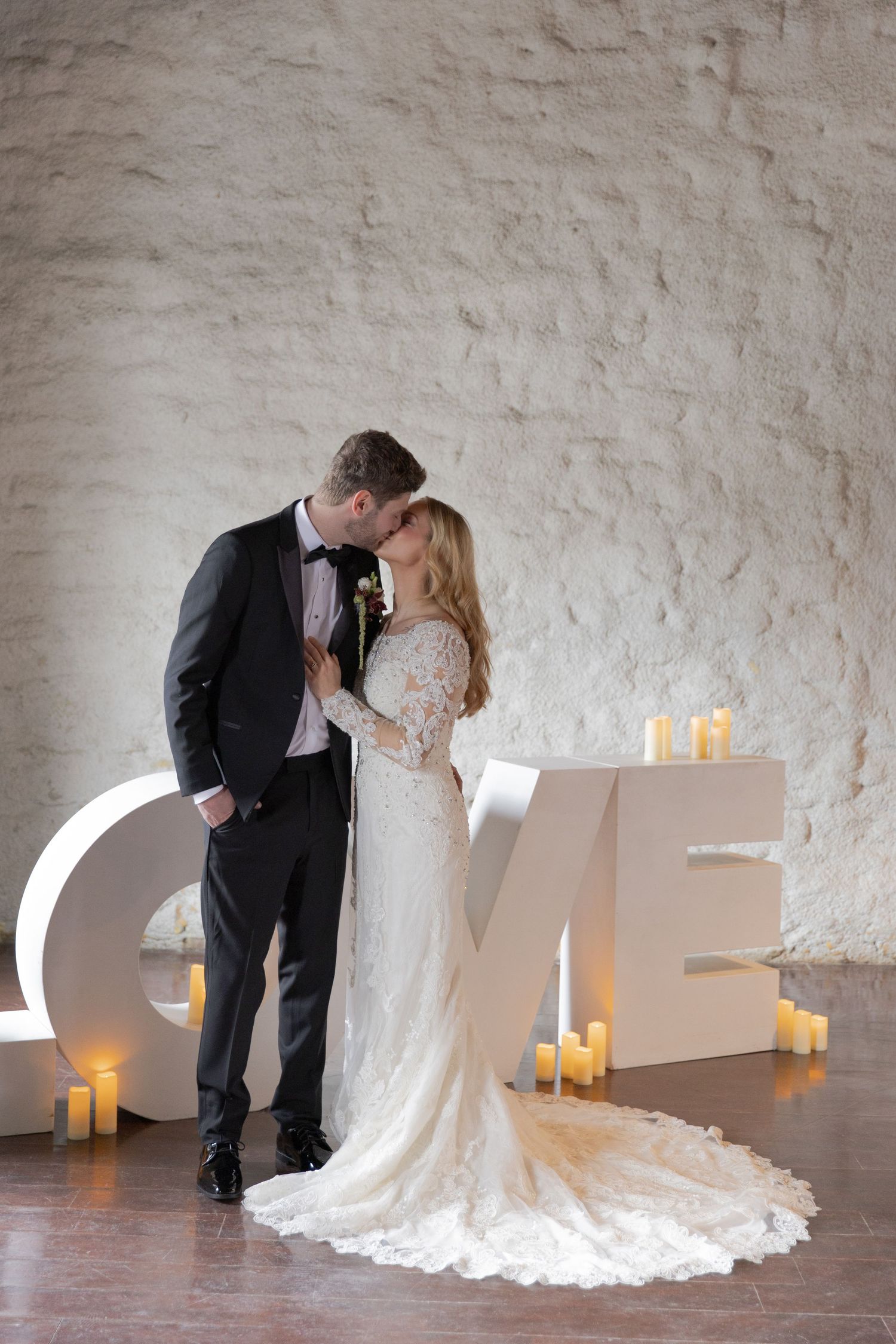 A romantic wedding couple stands by illuminated LOVE letters with candles against a textured white wall.