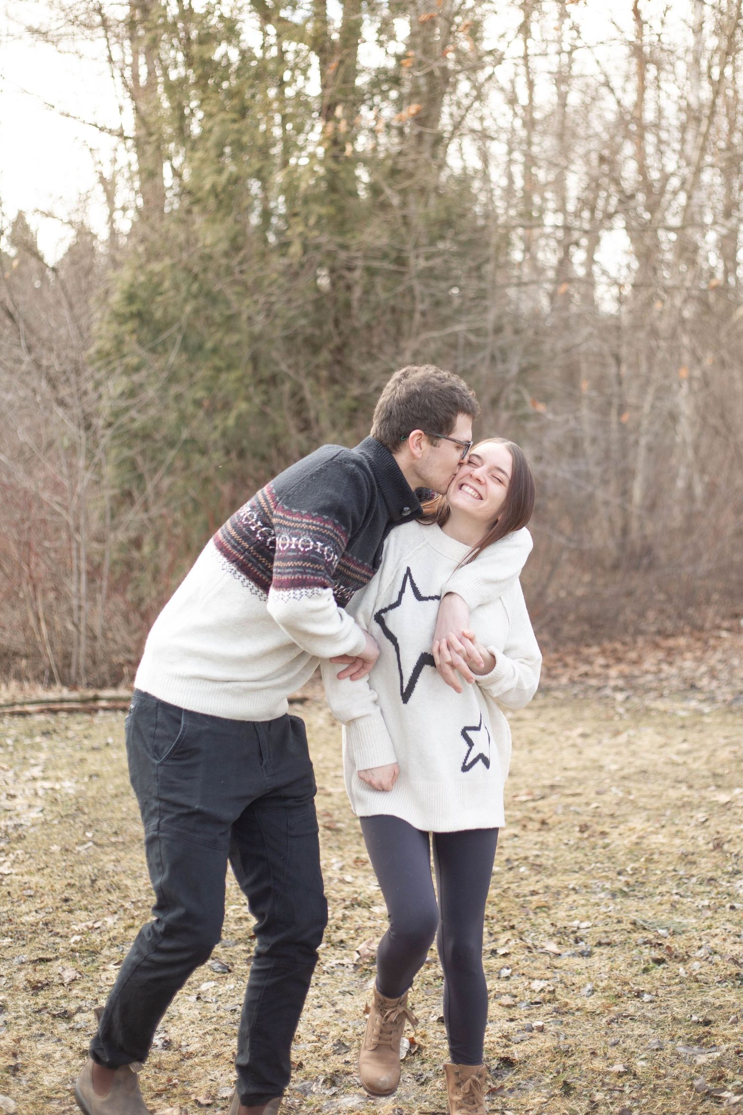 A couple shares a playful moment together outdoors in winter wearing cozy sweaters against a wooded background.