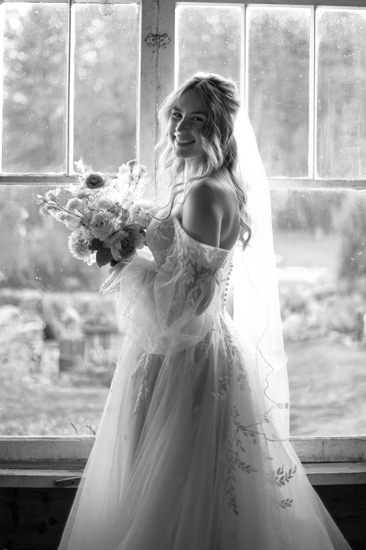 Black and white bridal portrait showing an off-shoulder lace wedding gown and flowing veil near a window.