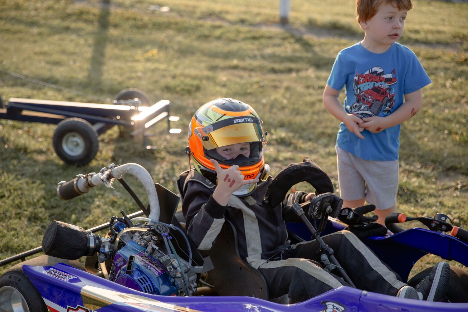 A racing go-kart with orange helmet and racing equipment parked on grass with a spectator nearby.