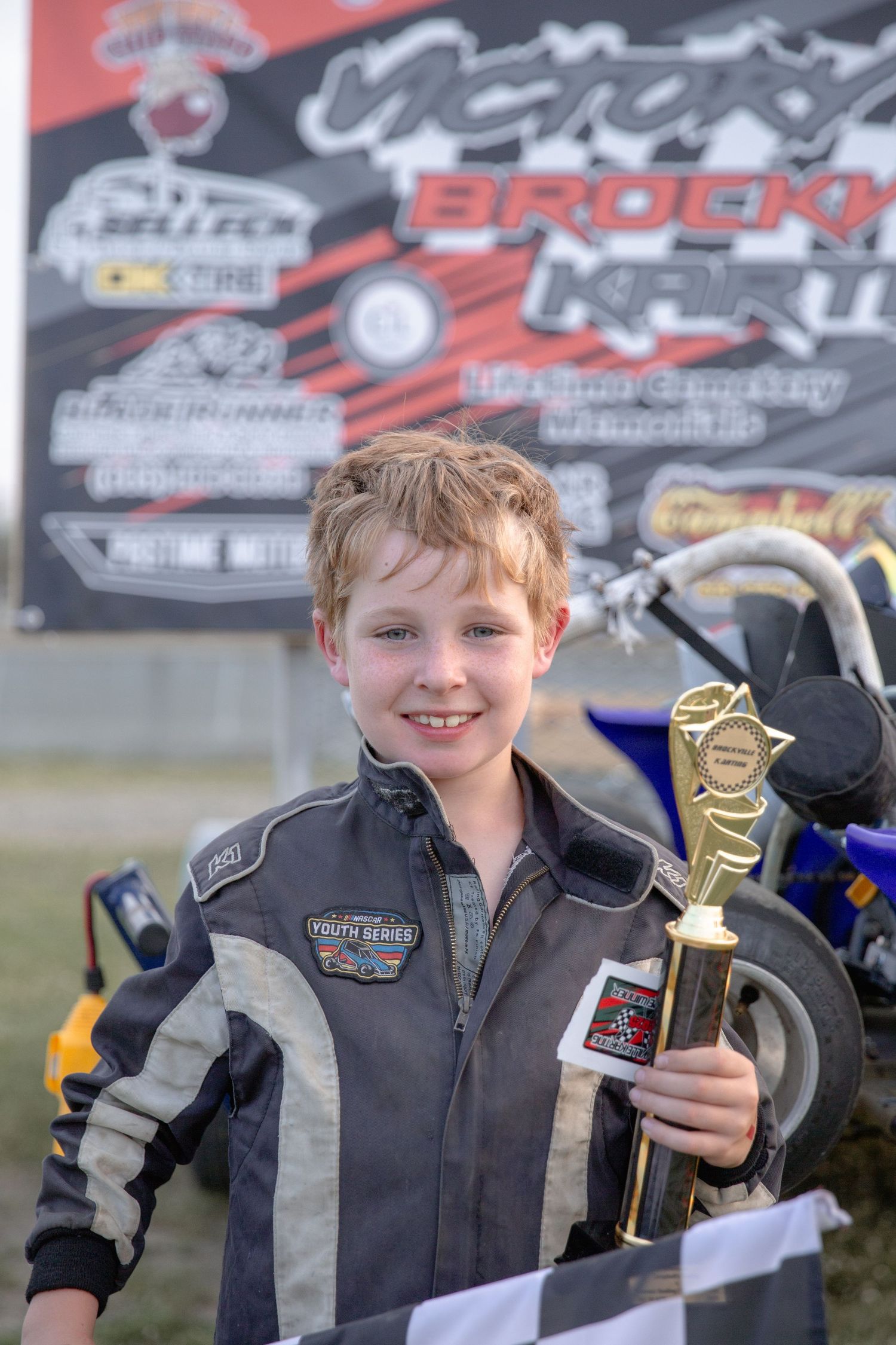 Young racing champion proudly displays multiple trophy awards at motorsports event with racing banners in background.