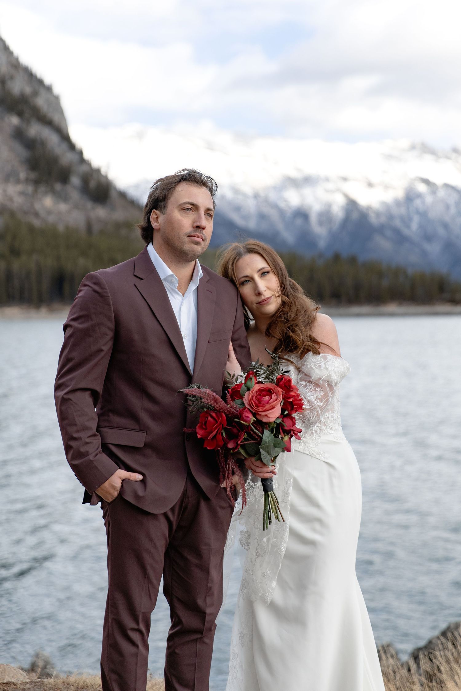 Newlyweds in burgundy and white pose together against a dramatic mountain lake backdrop.