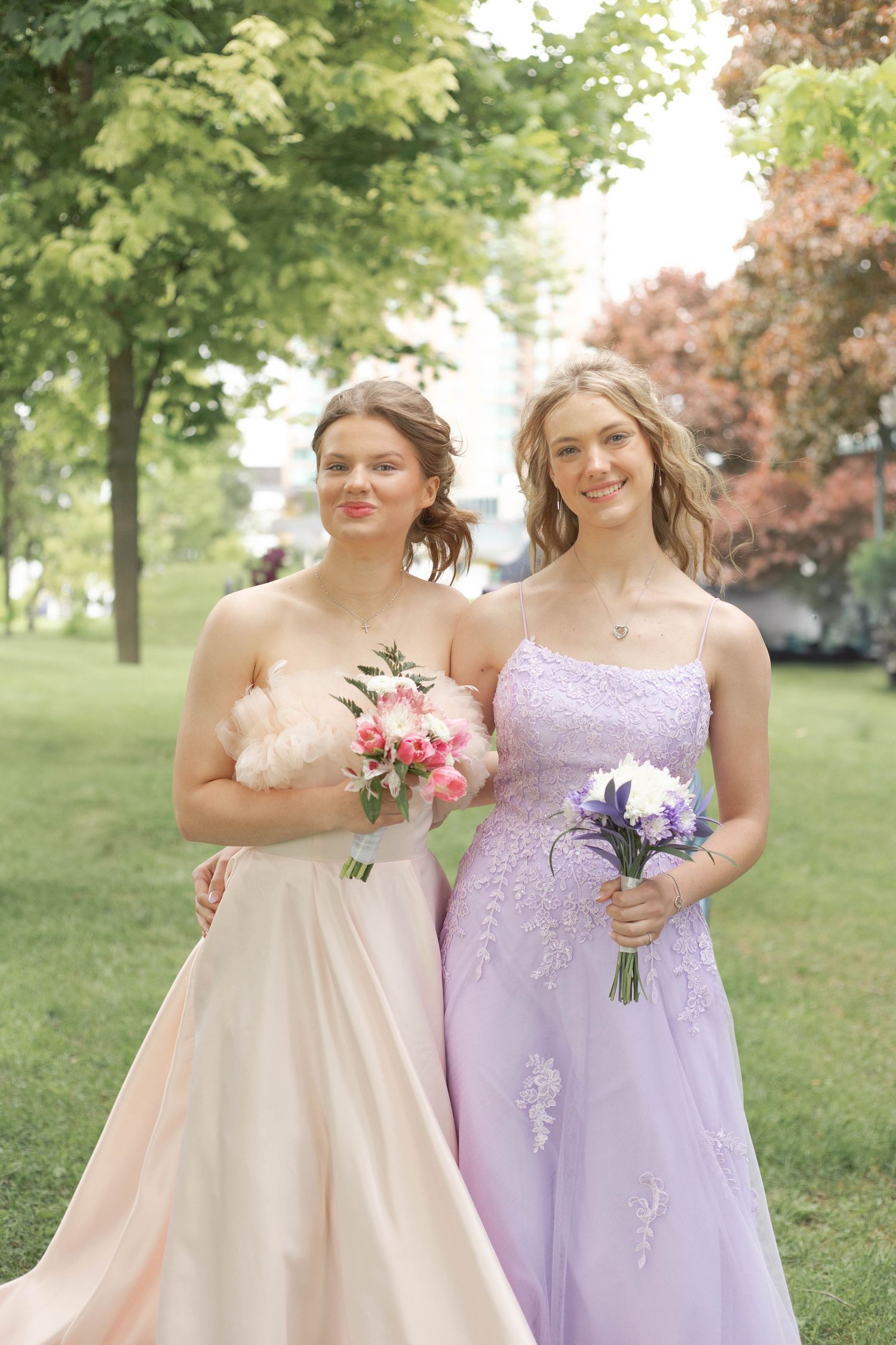 Two bridesmaids in pink and purple strapless gowns hold floral bouquets while standing in a garden setting.
