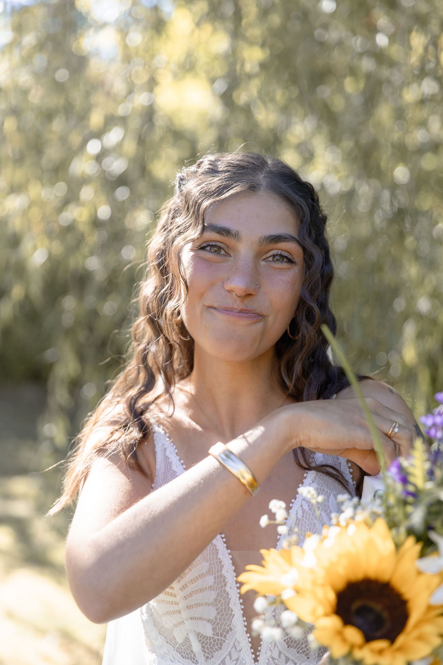 A person in a white lace dress poses playfully with bright yellow sunflowers in a sunny outdoor setting.