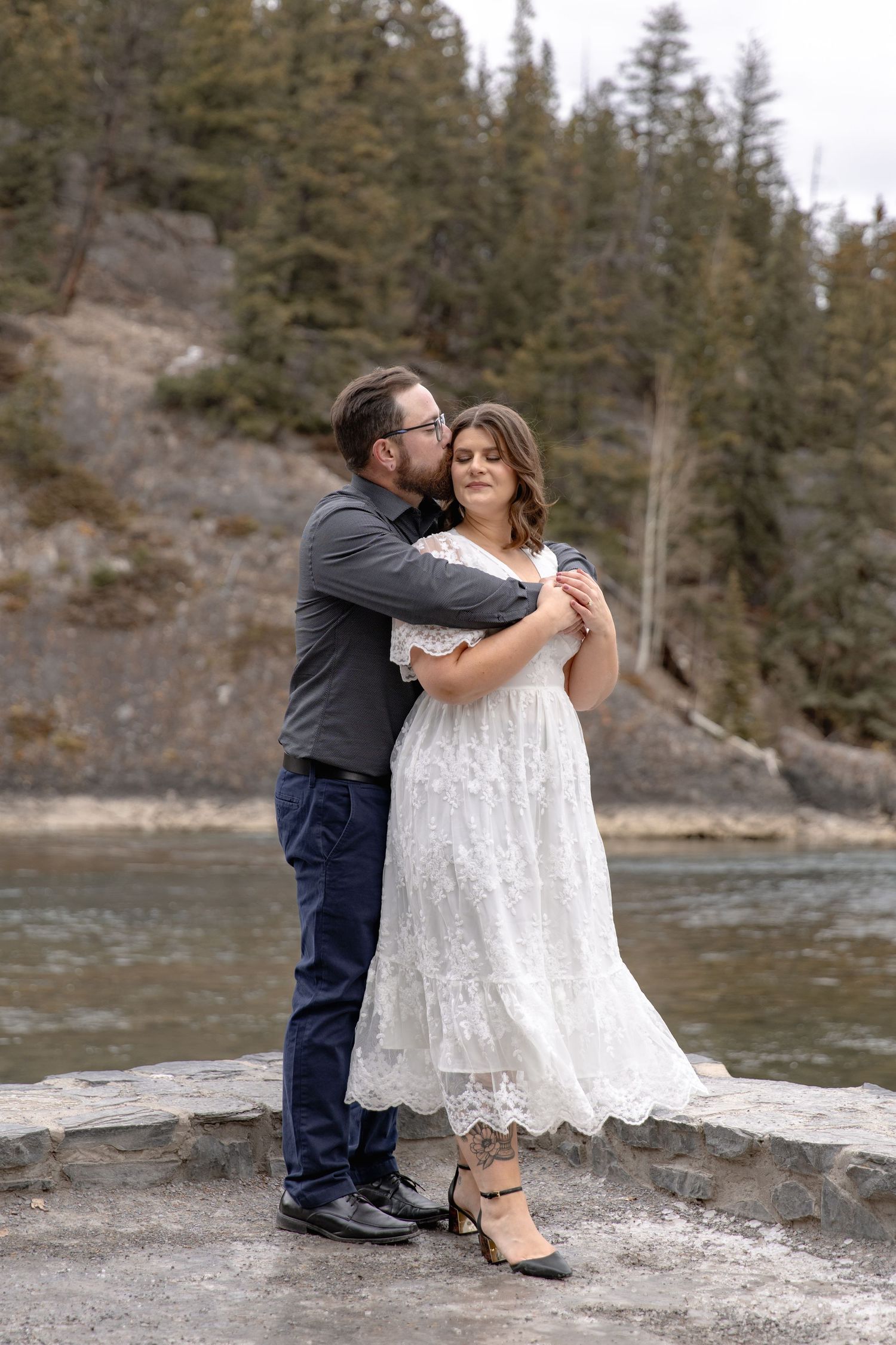 A couple shares an intimate moment on rocky riverbank surrounded by evergreen forest.